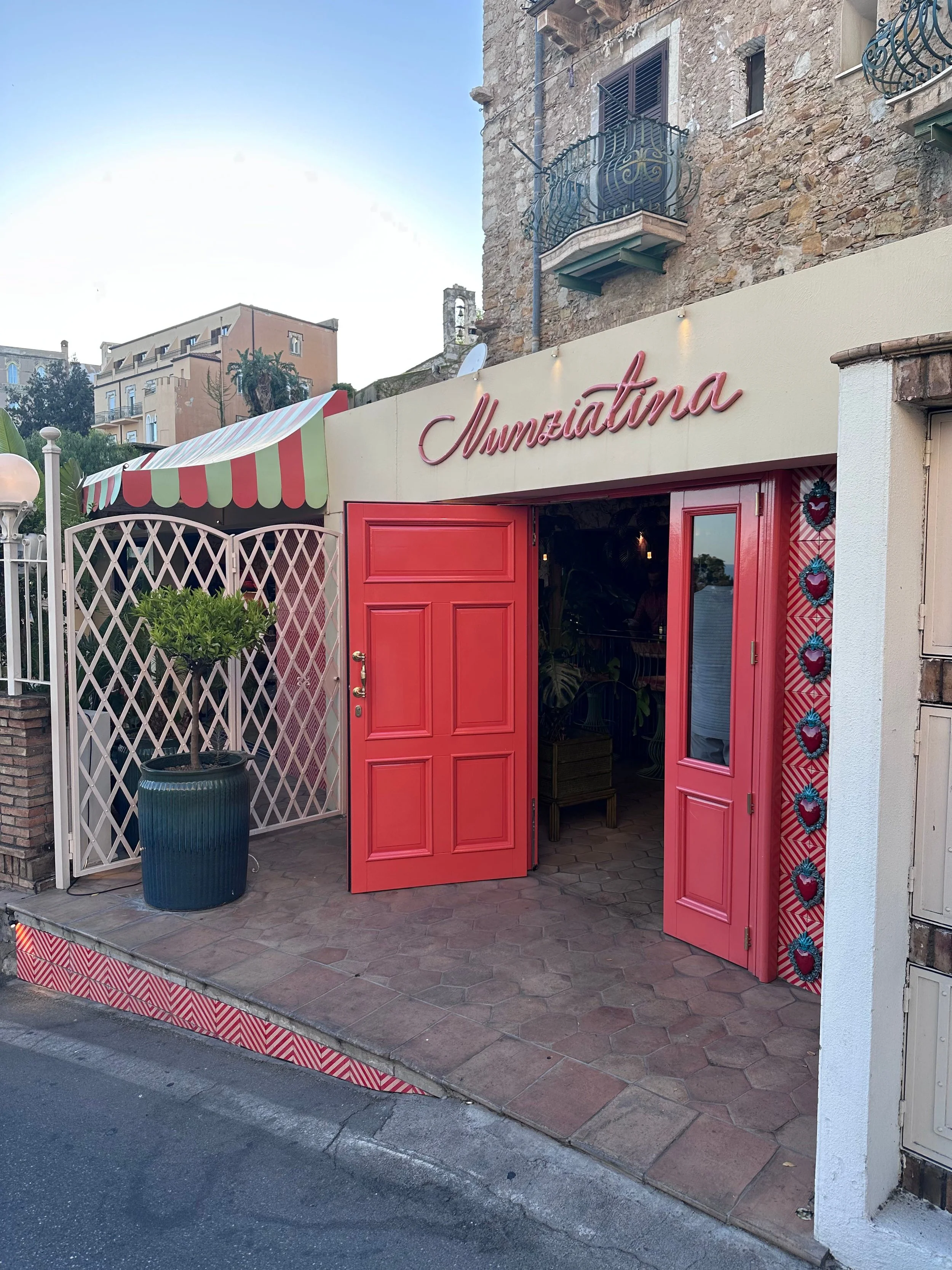 Entrance to Numziatina restaurant with vibrant red door, potted plant, and a striped canopy beside an old stone building.