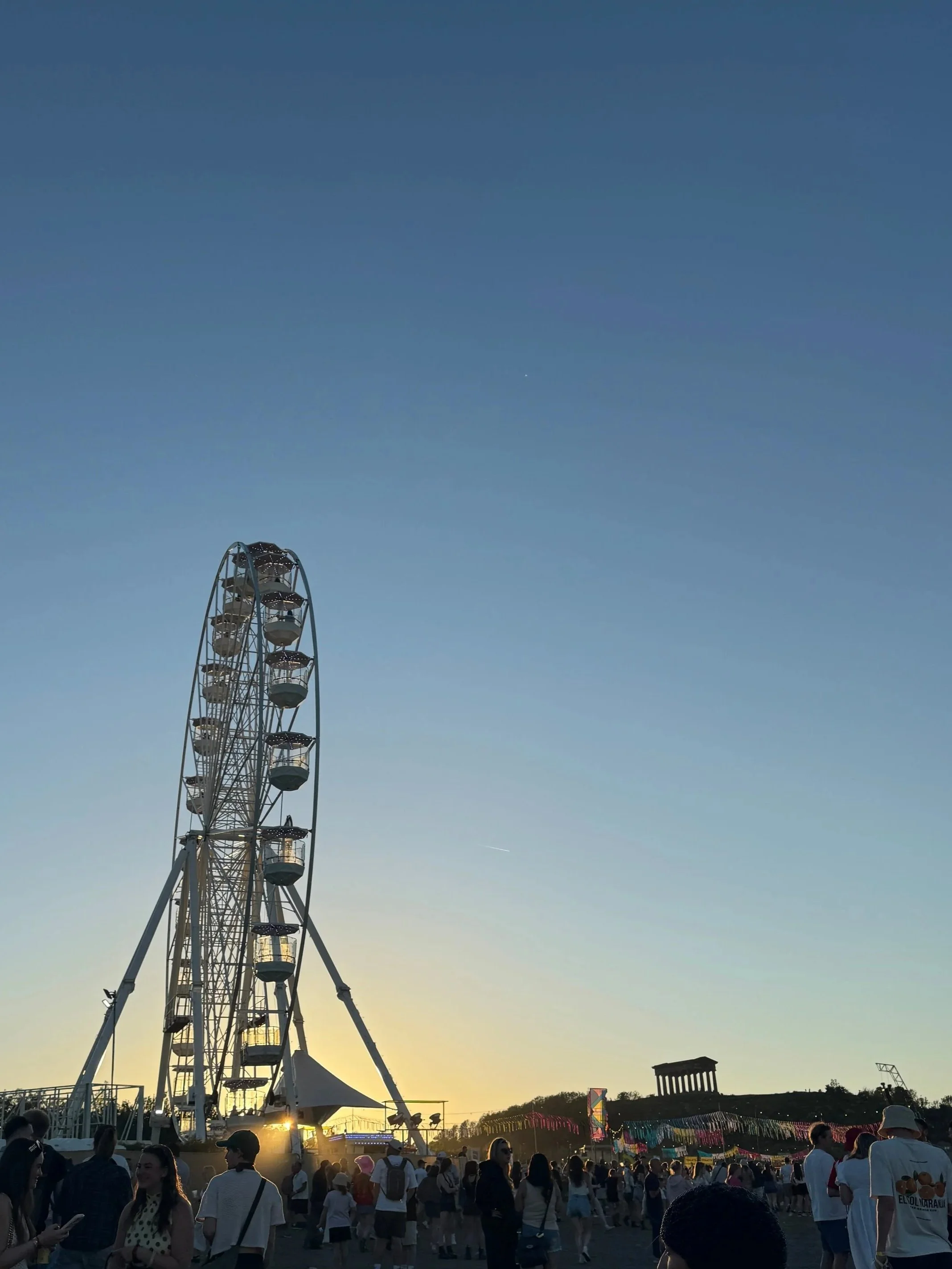 People stand near a ferris wheel, enjoying the warm hues of sunset as the sky transitions to evening.
