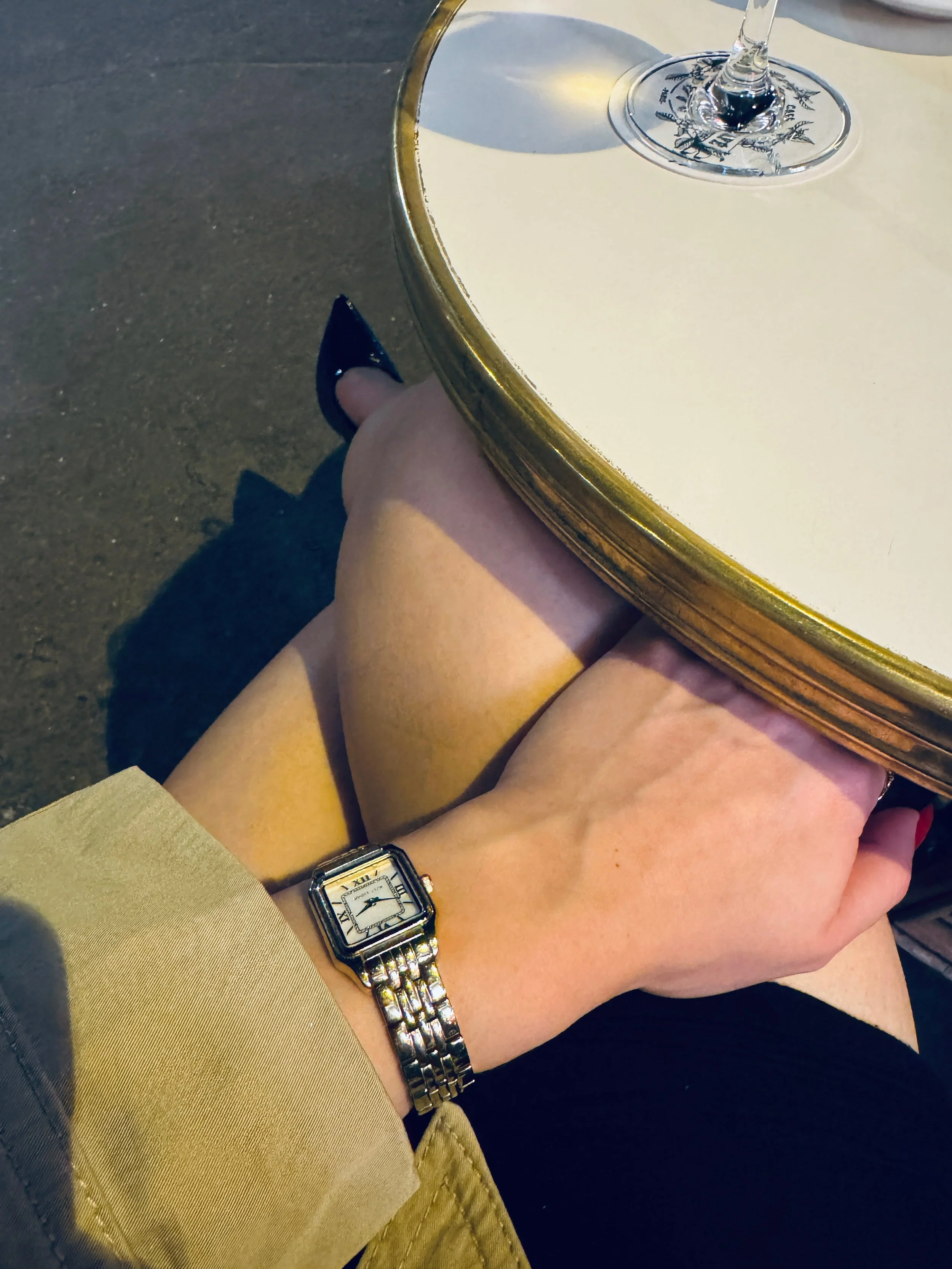Moonlit close-up of crossed legs and a hand beneath a round white café table, wearing a gold rectangular watch; a wine glass rests above.