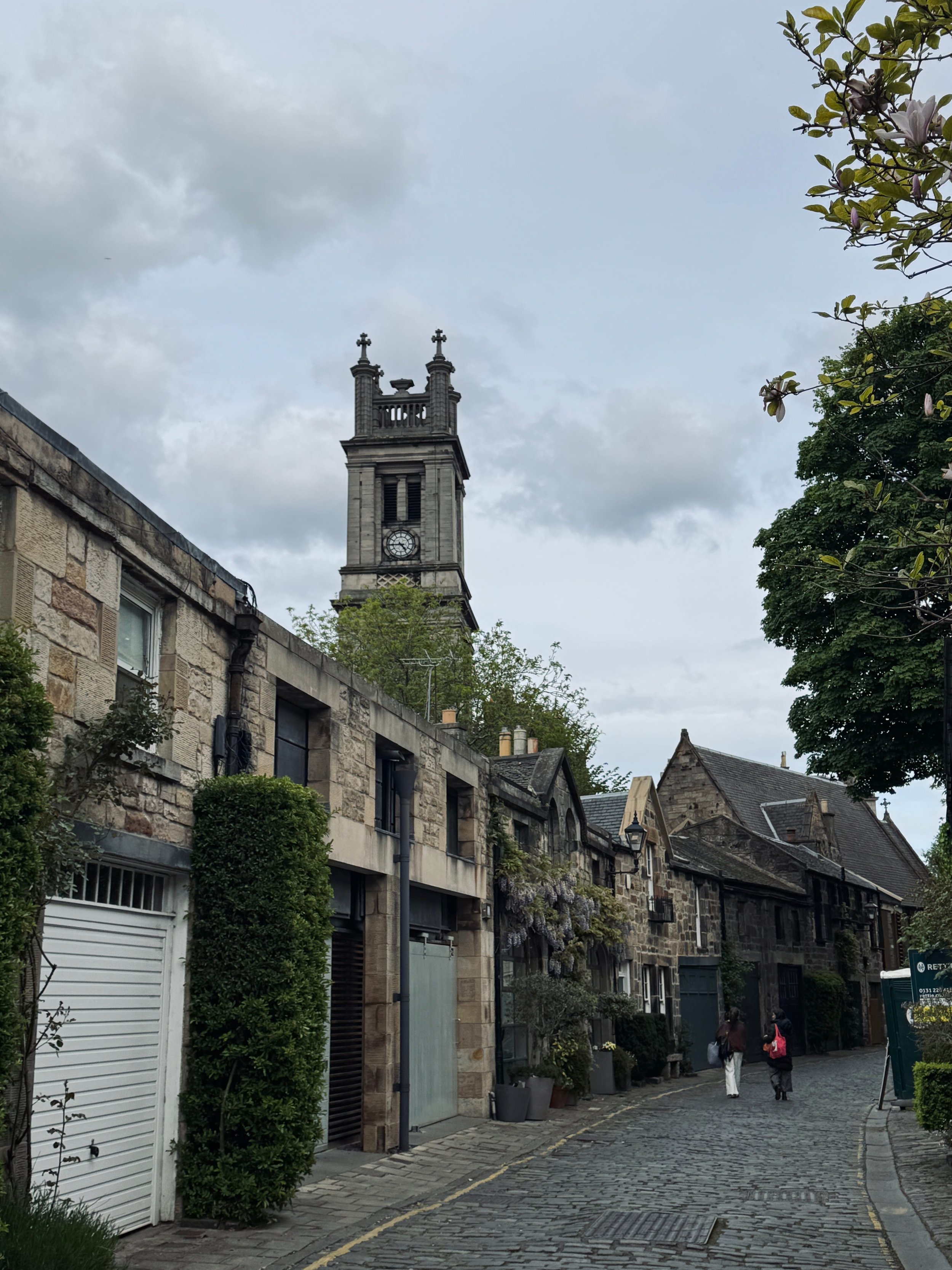 Narrow cobblestone street with stone buildings and lush greenery, leading to a tall clock tower under a cloudy sky.
