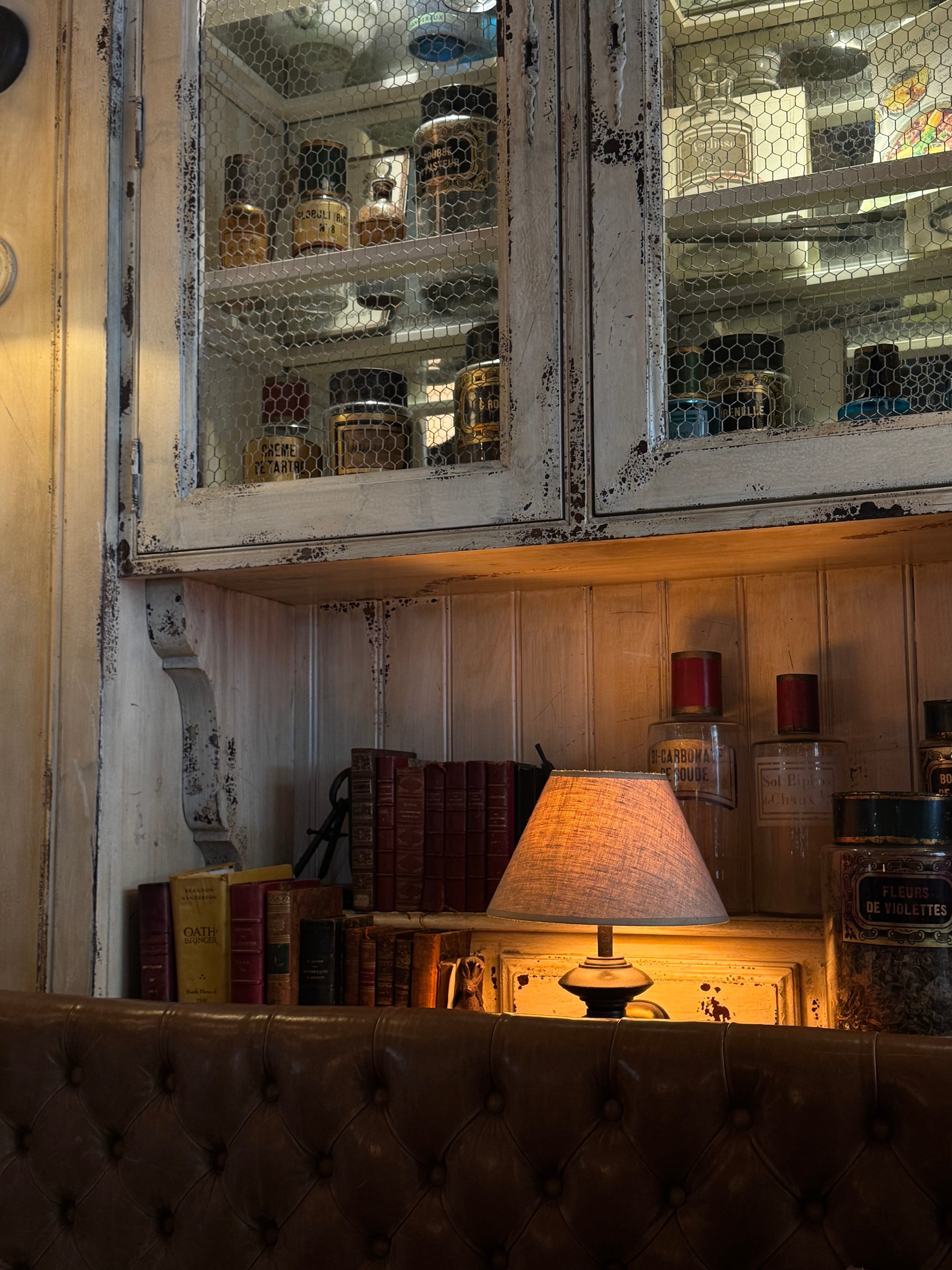 A warm table lamp glows among old books beneath a distressed glass-front cabinet of jars, with a dark tufted leather chair creating a cozy vintage mood.