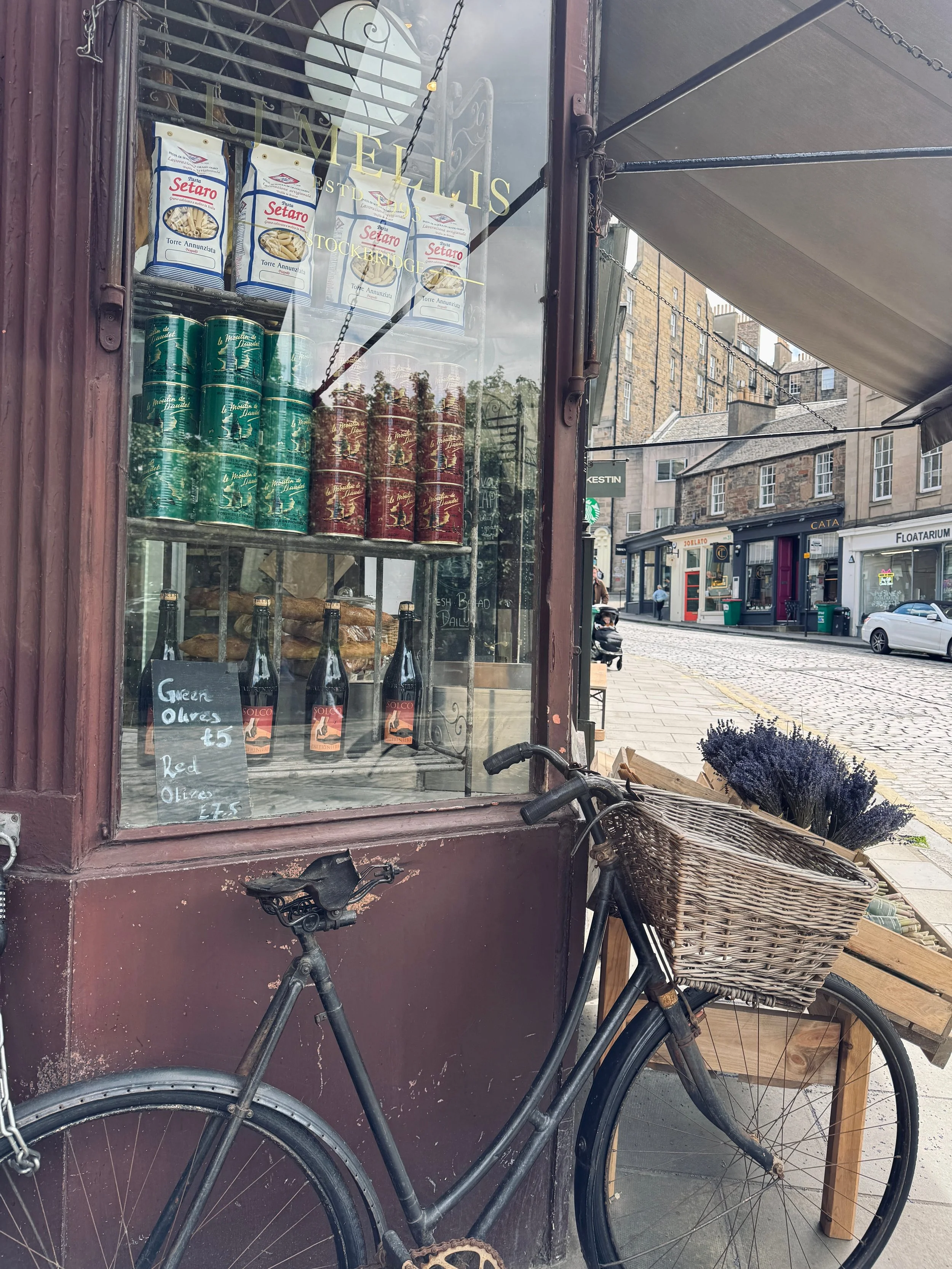 Old bicycle with basket of lavender leans against a deli window displaying pasta, tins, and wine bottles. Cobblestone street in background. Cozy, rustic vibe.