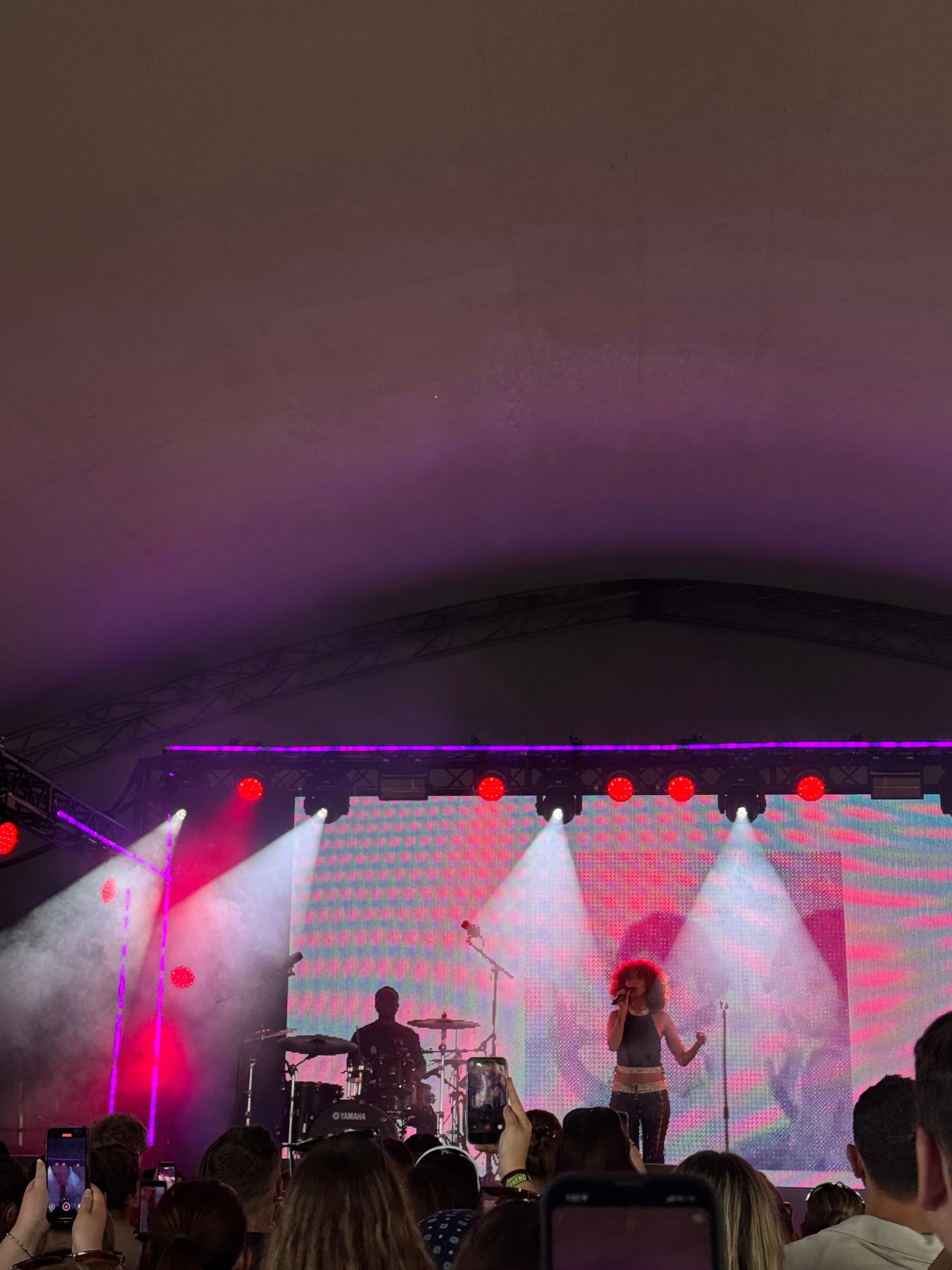 Live concert with a female singer and drummer onstage, surrounded by pink and purple lights, smoke, and a large colorful LED screen; crowd watches.
