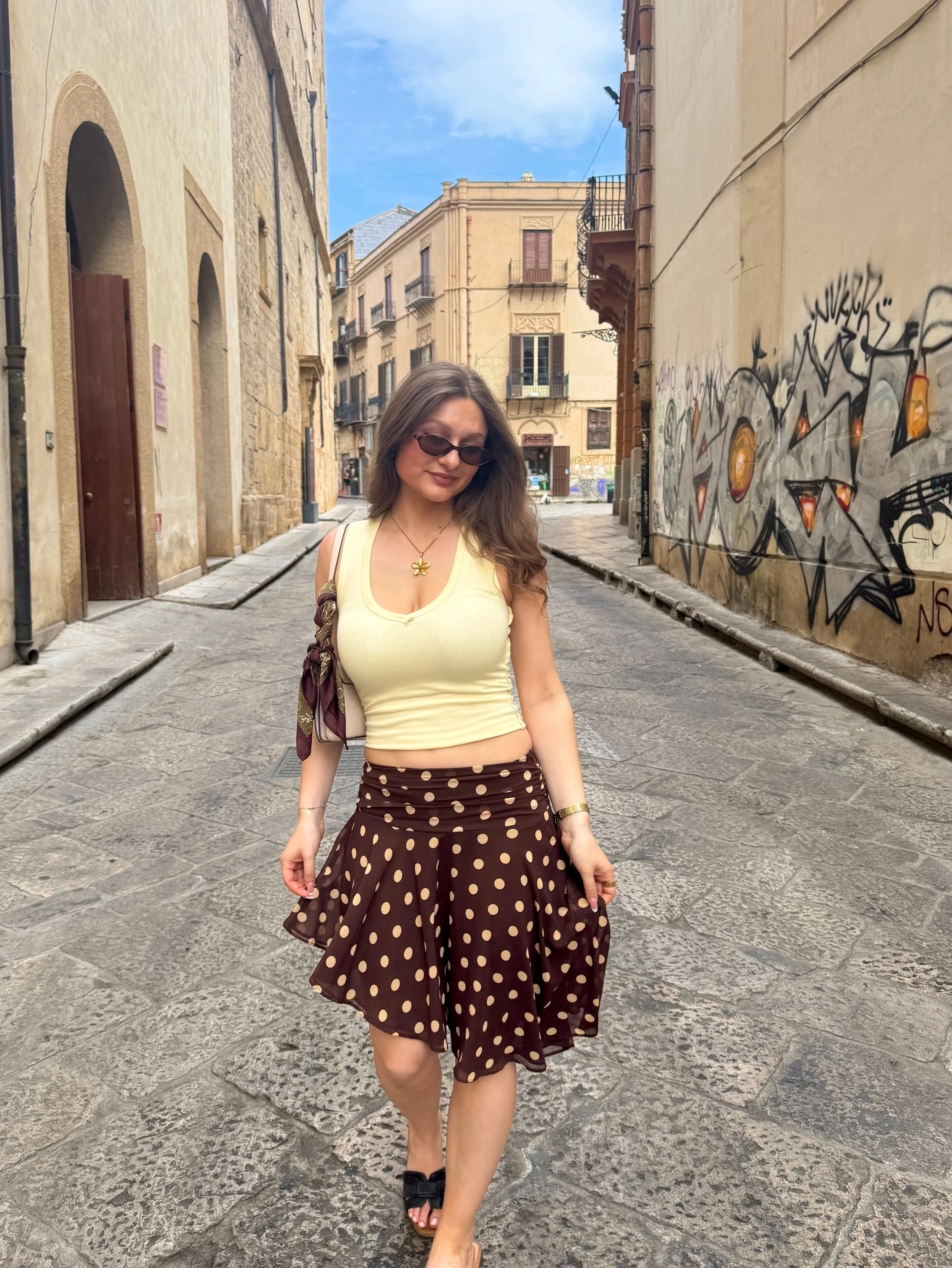 A woman in a polka dot skirt and yellow top strolls confidently down a vibrant alleyway.
