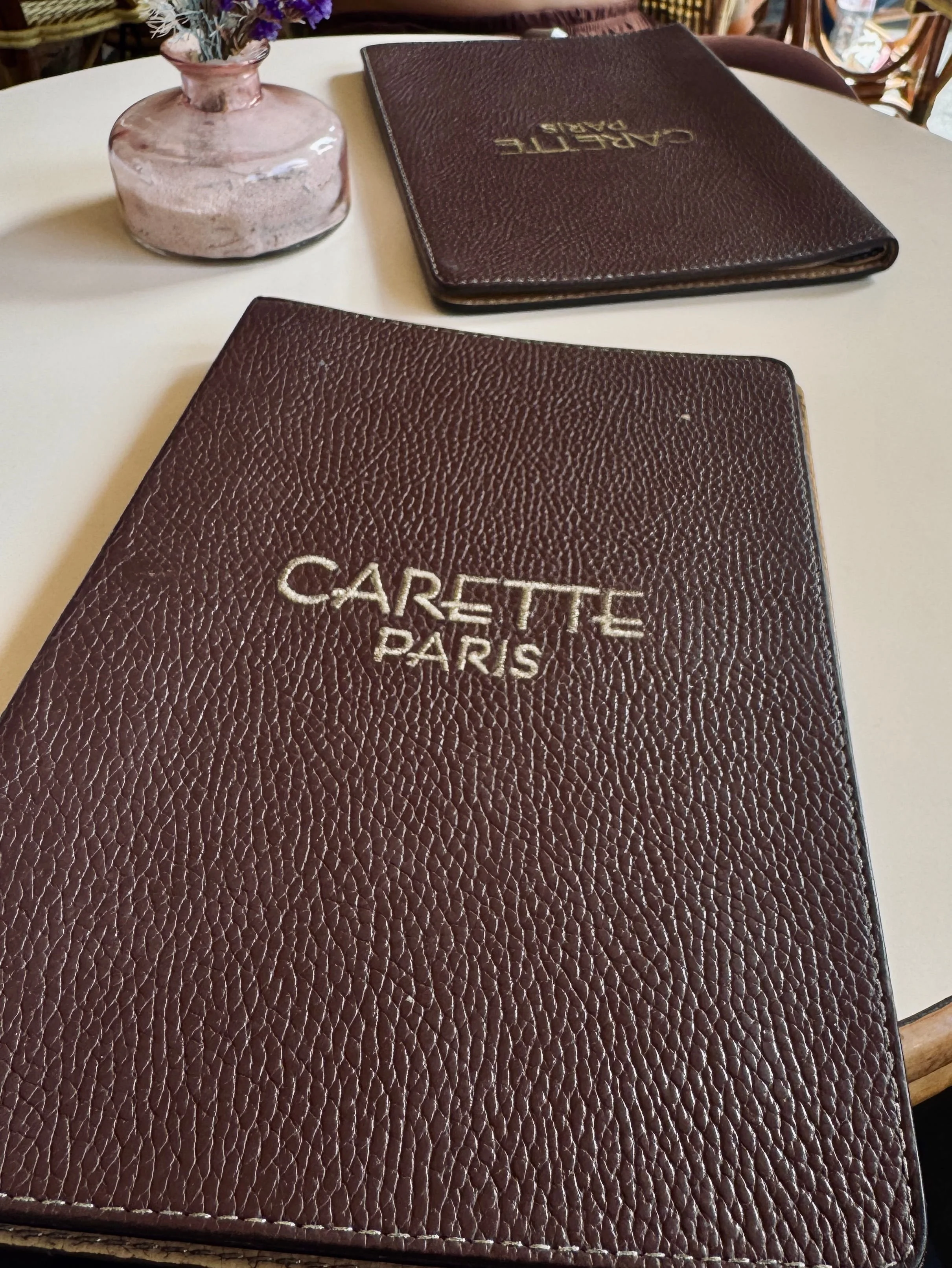 Two dark brown, textured Carette Paris menus rest on a cream café table beside a small pink vase, creating an elegant, intimate dining atmosphere.