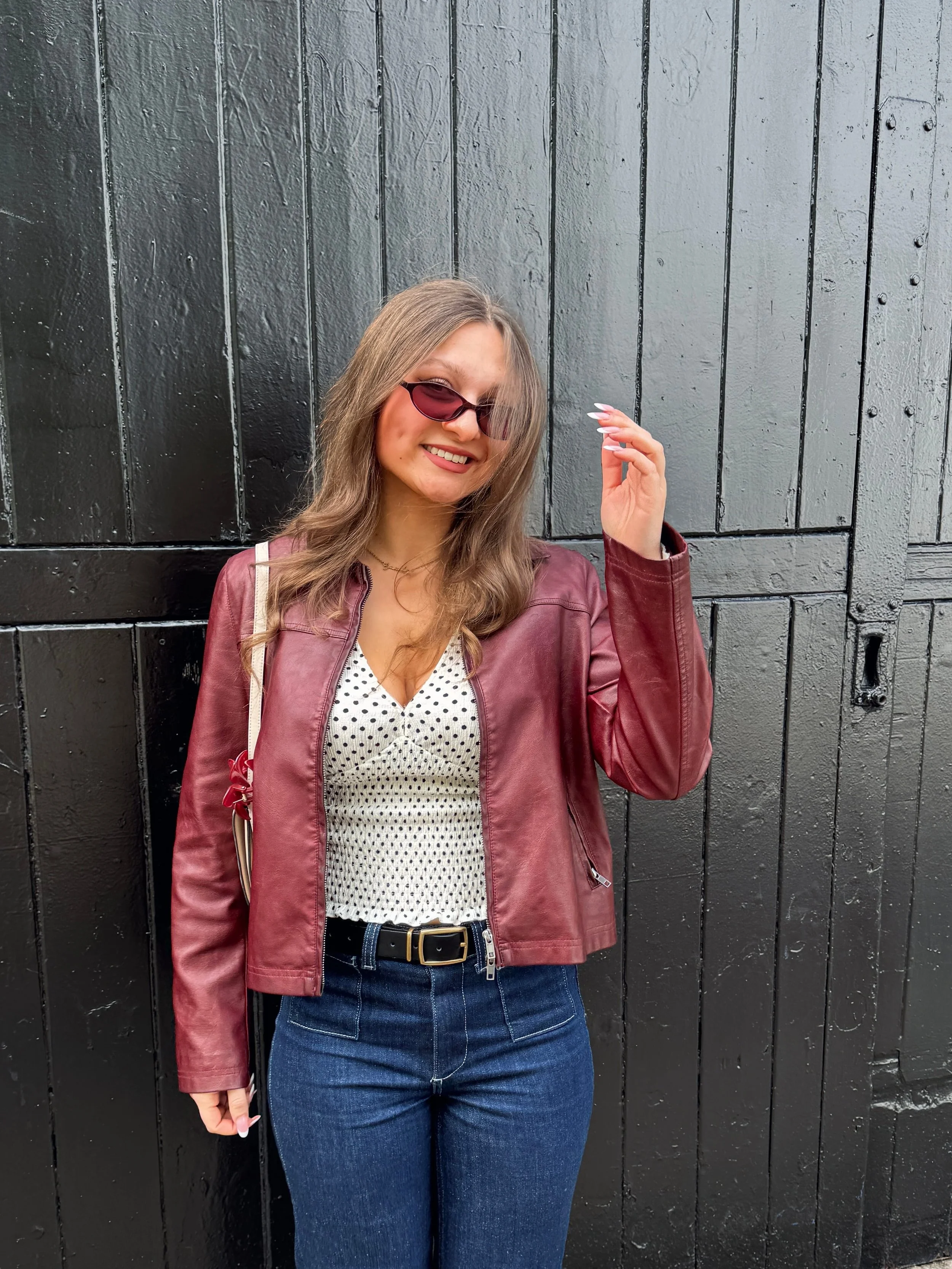 A woman smiling in front of a black wooden door, wearing a maroon jacket, polka dot top, and jeans. She has sunglasses and a casual pose.