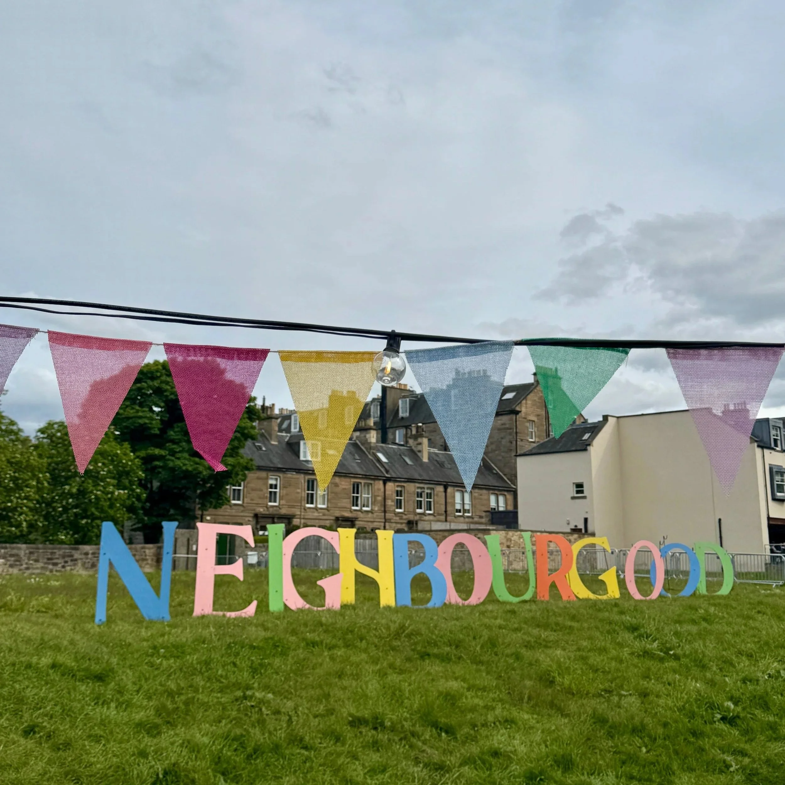 Colorful bunting hangs above large, multicolored letters spelling "NEIGHBOURGOOD" on a grassy field, with buildings and cloudy sky in the background.