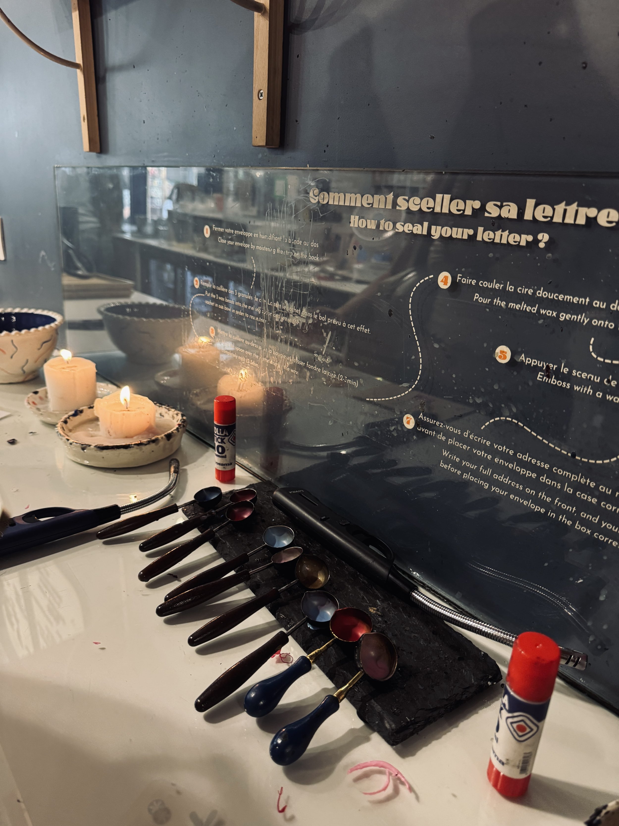 Letter-sealing station with candles, red wax sticks, and dark-handled tools beneath French-English instructions; reflective, antique atmosphere.
