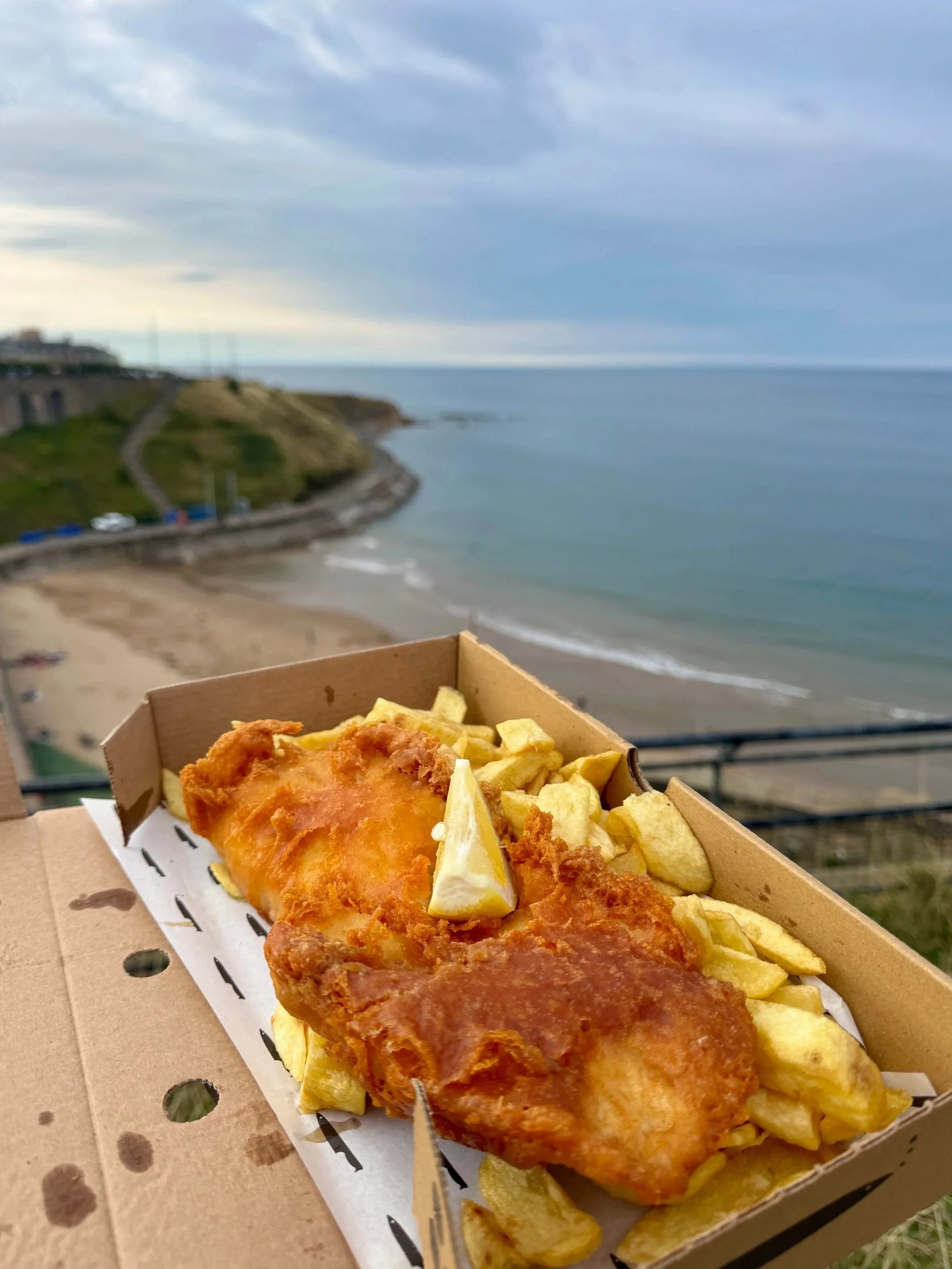 Golden battered fish and chips in a cardboard takeaway box overlooking a quiet beach, sea, and cloudy coastal horizon; calm seaside mood.