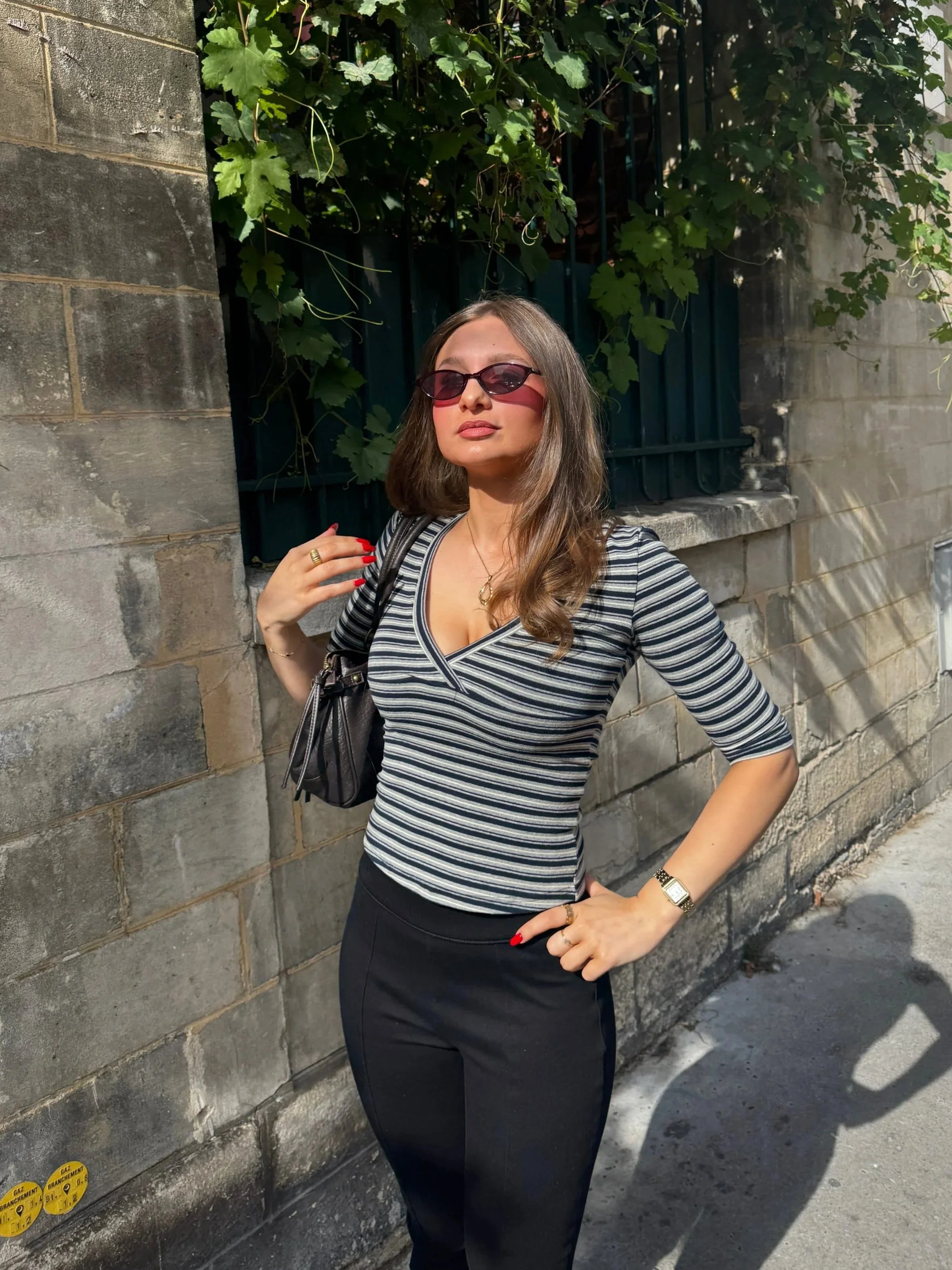 Woman in sunglasses poses confidently by a vine-covered stone wall, wearing a striped top and black pants in bright sunlight.