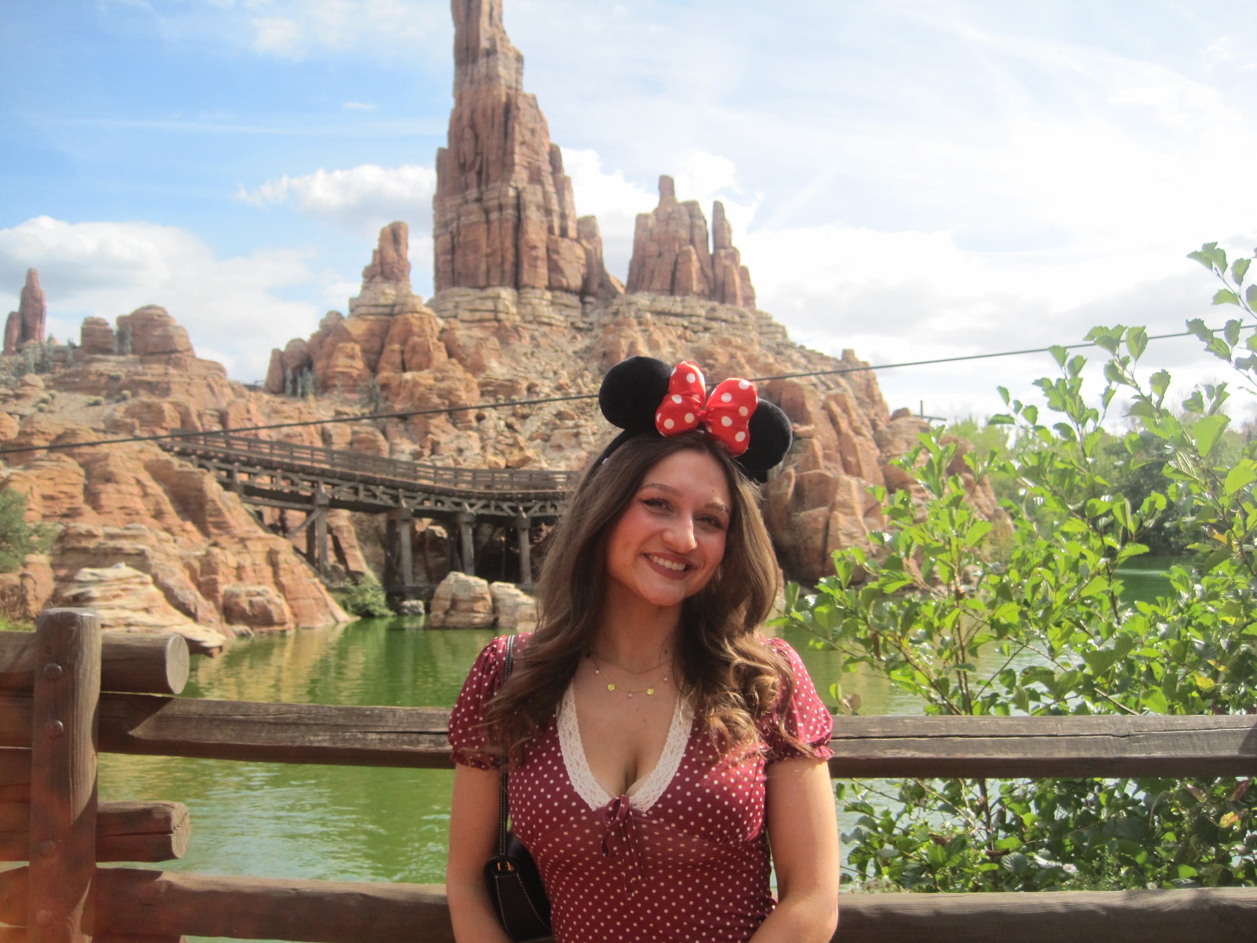 A smiling woman wearing Minnie Mouse ears and a red polka-dot dress poses by a lagoon, with dramatic desert rock spires behind her. The scene feels cheerful and sunny.