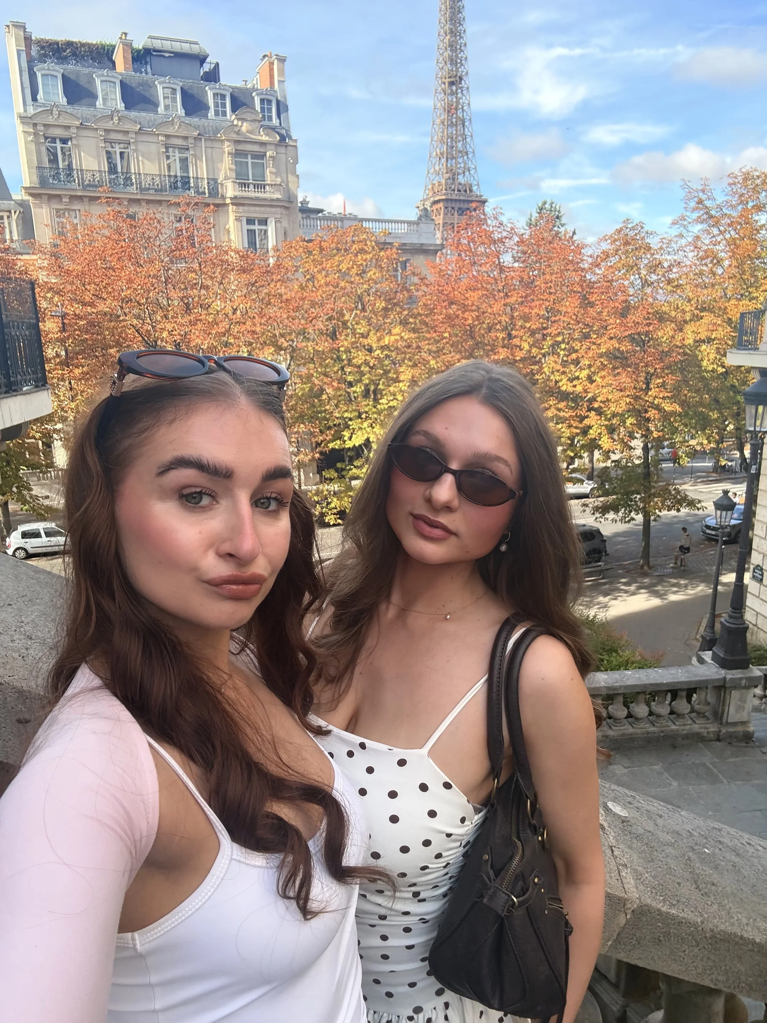 Two women pose by orange autumn trees in Paris, with an ornate historic building and the Eiffel Tower behind them under a blue sky; relaxed, stylish mood.
