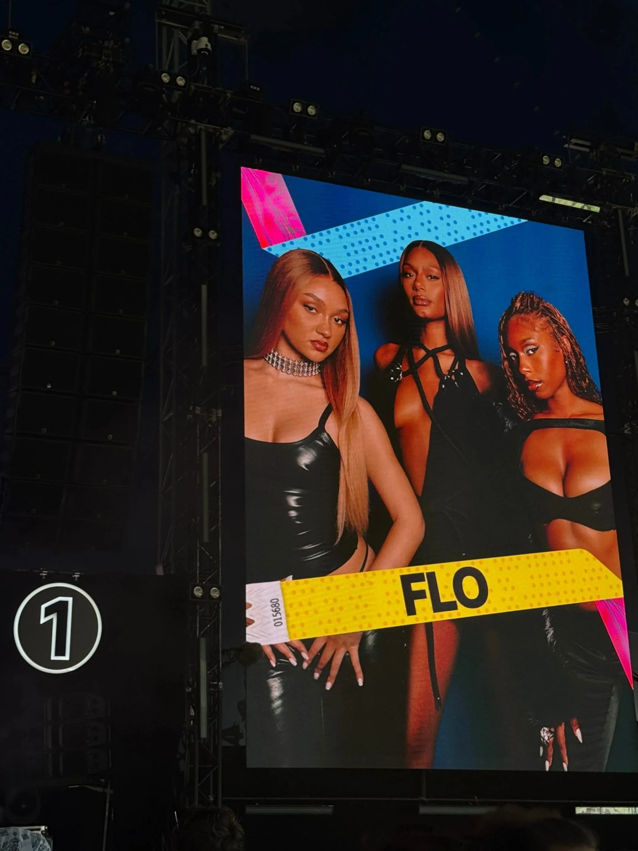 Large outdoor screen at night showing three women posing, with a yellow banner labeled "FLO" across the bottom.