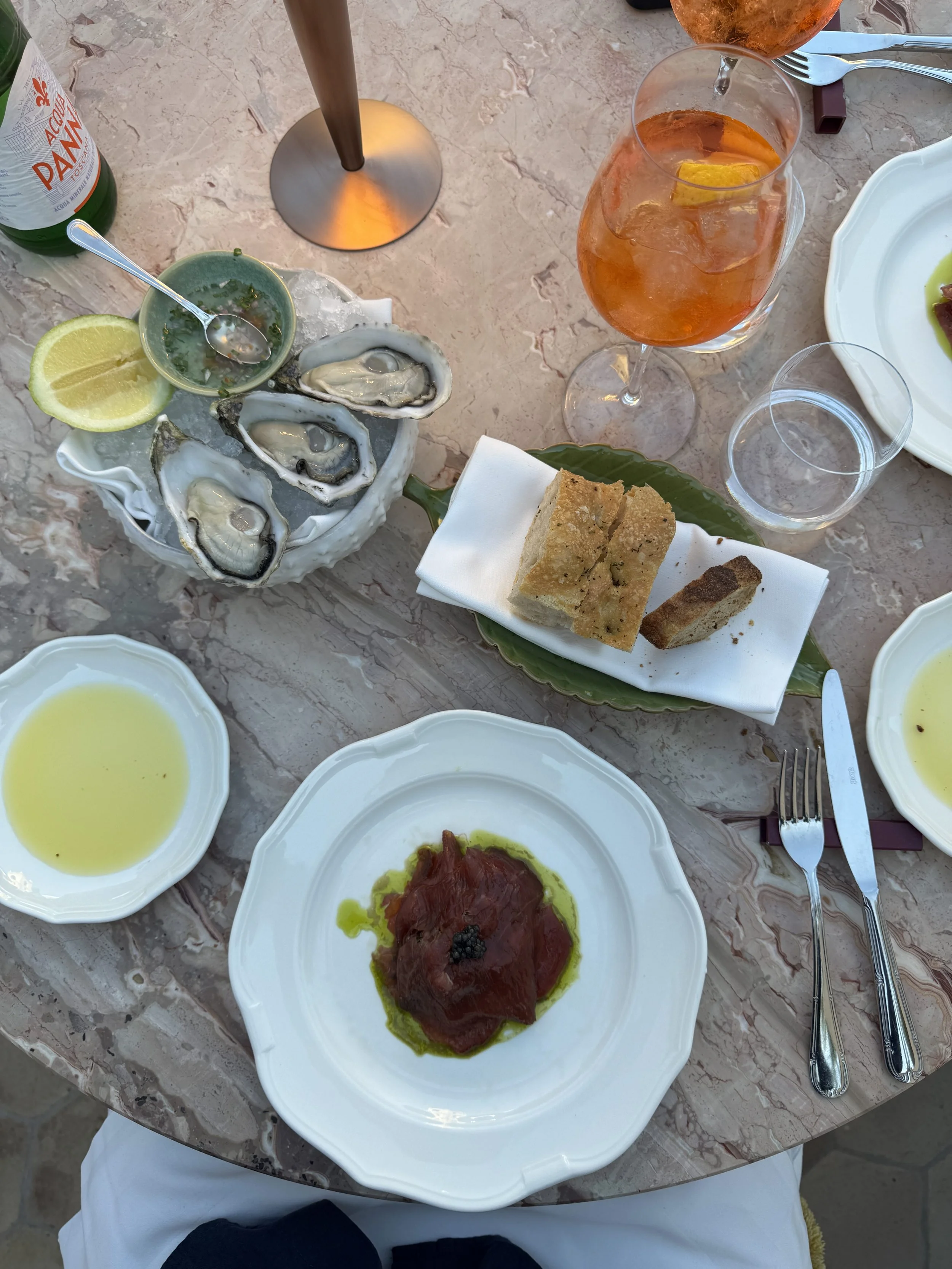 A table setting with a marble surface featuring plates of oysters, a dish with sliced fish, bread, and a cocktail.