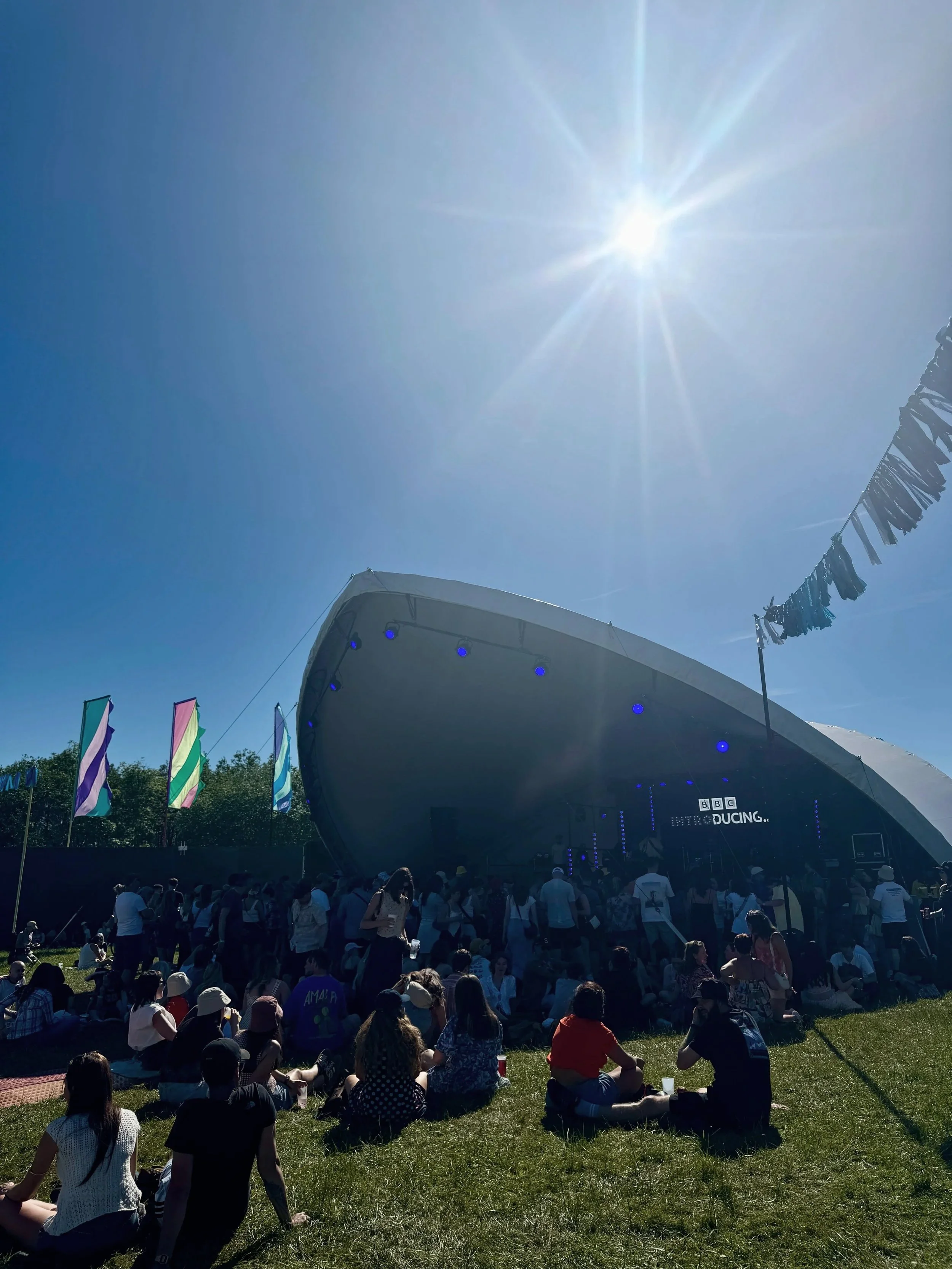 Crowd enjoying a sunny outdoor music event near a BBC Introducing stage with colorful flags and blue sky overhead.