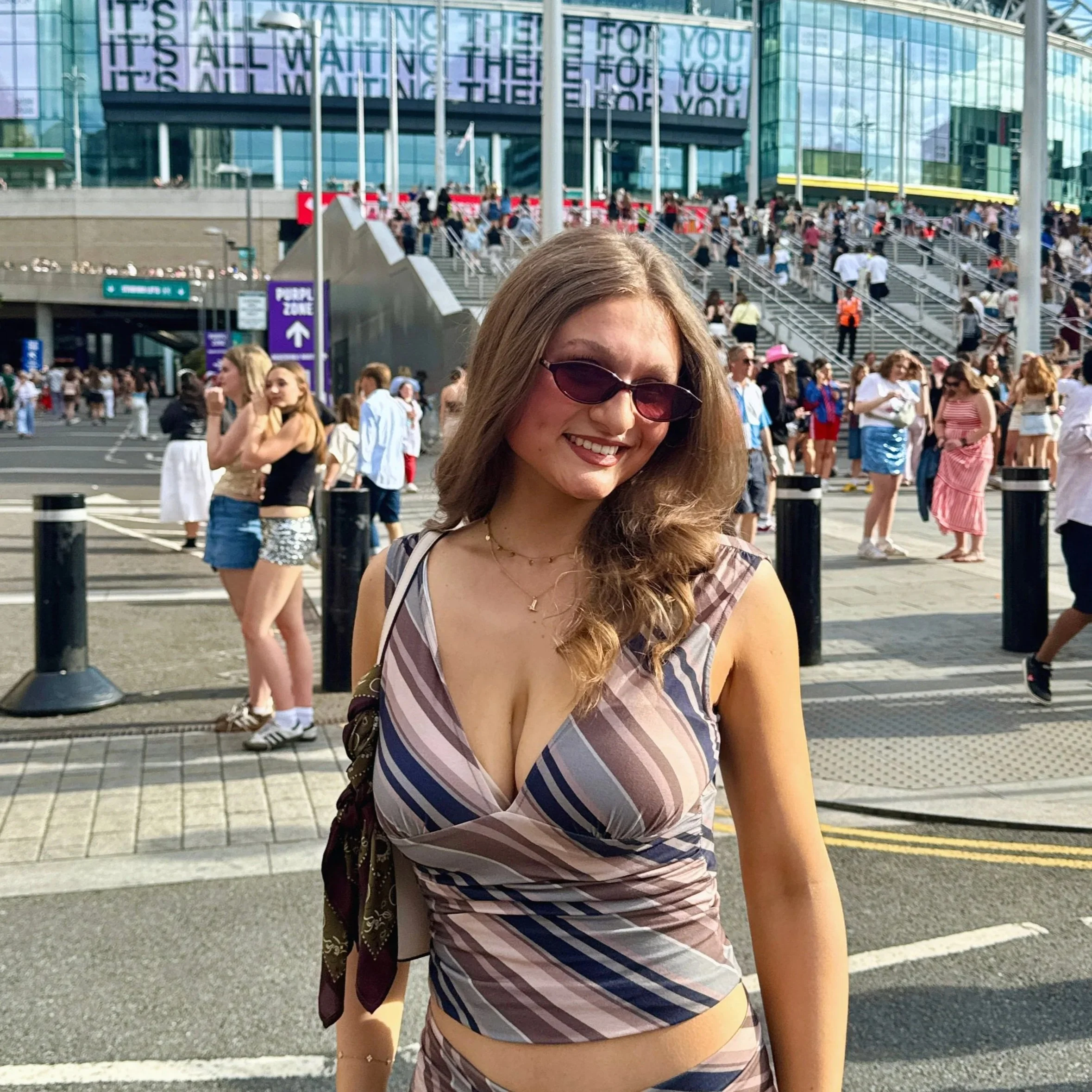 A woman in a striped outfit stands smiling outside Wembley stadium for the Harry Styles concert.