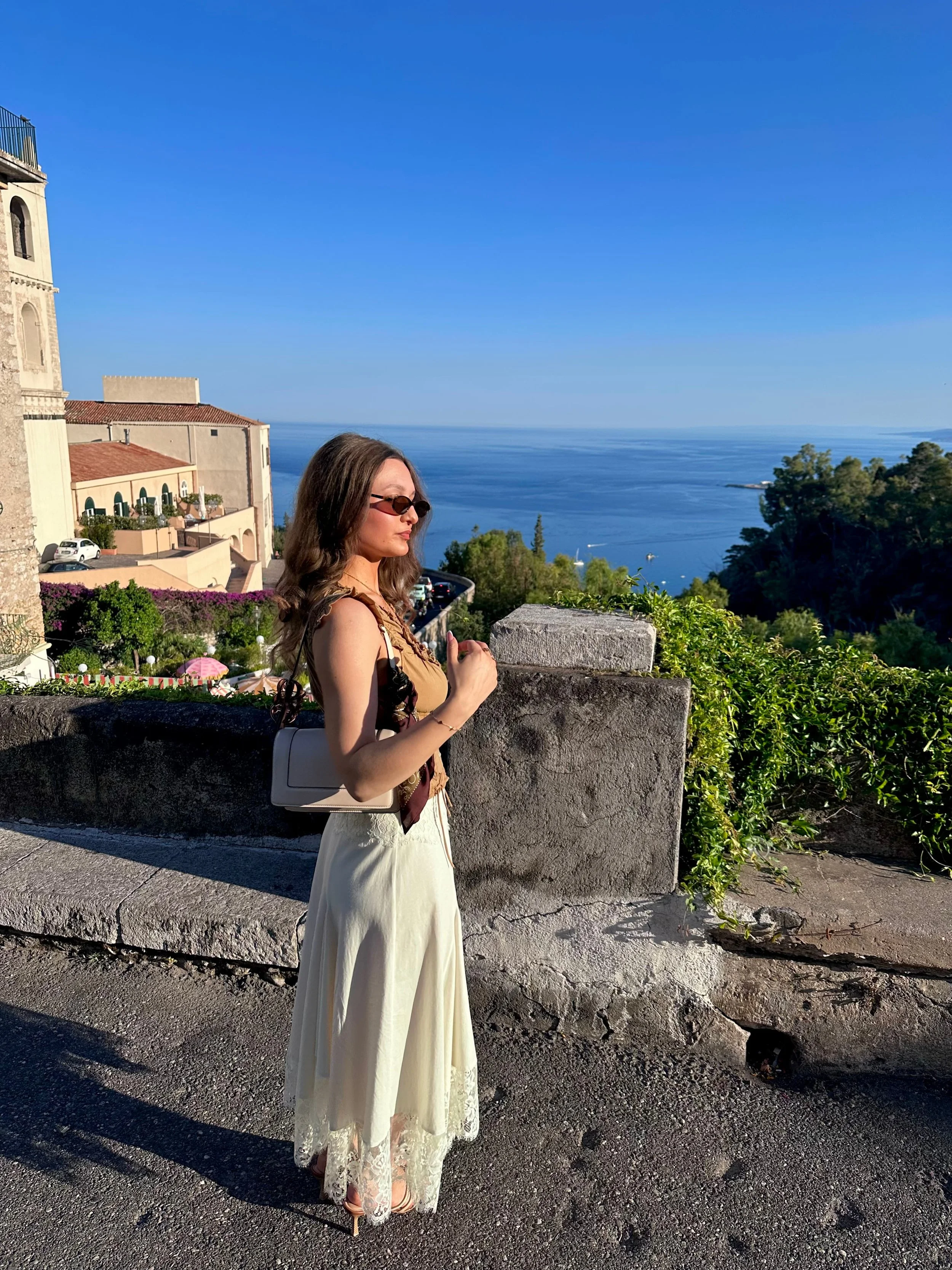 Woman in a beige top and white skirt stands on a sunny coastal street overlooking blue sea, greenery, and historic buildings.