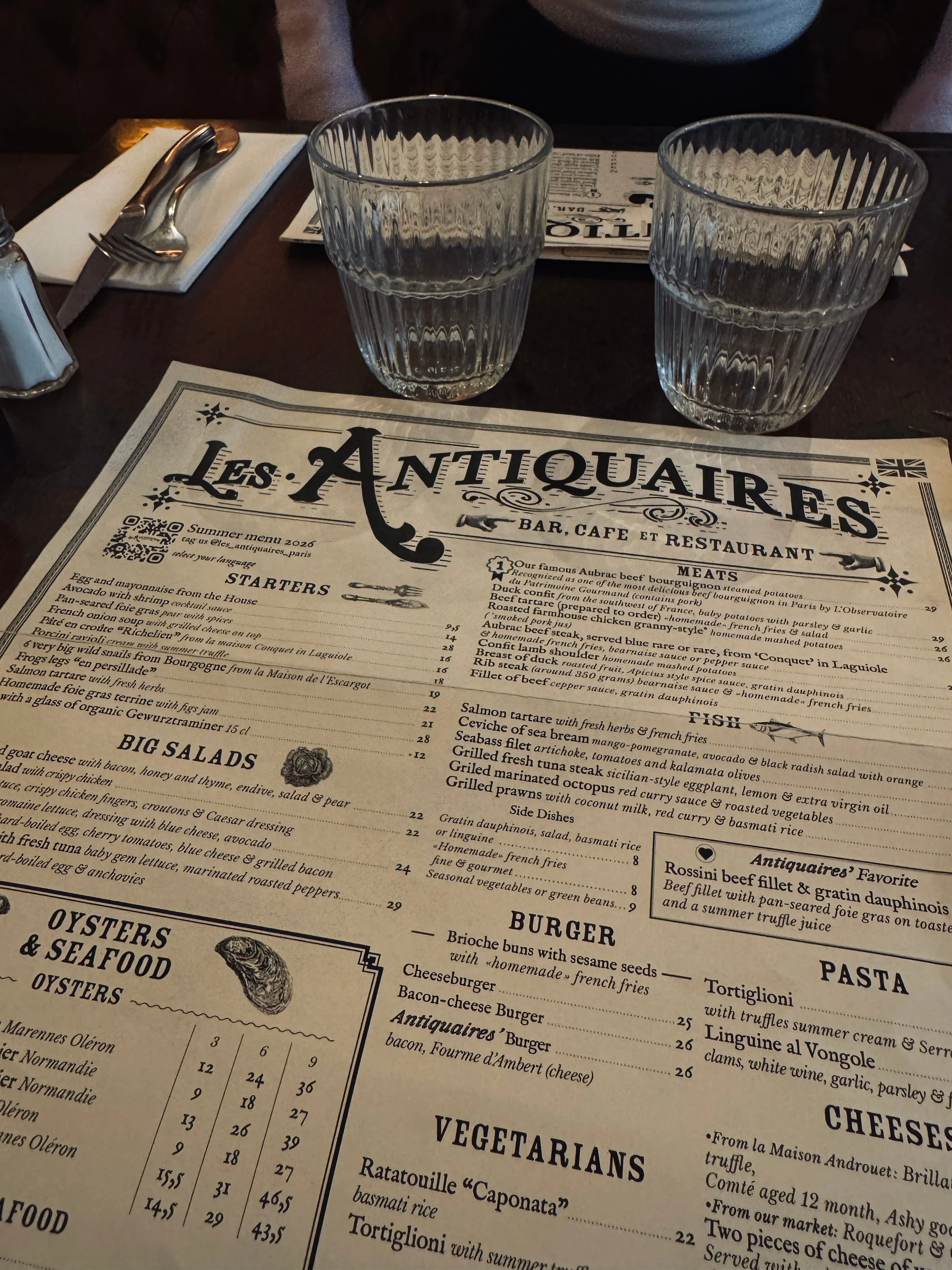 Vintage “Les Antiquaires” restaurant menu on a dark table, with two ribbed water glasses and silverware; its antique typography gives a cozy, elegant feel.