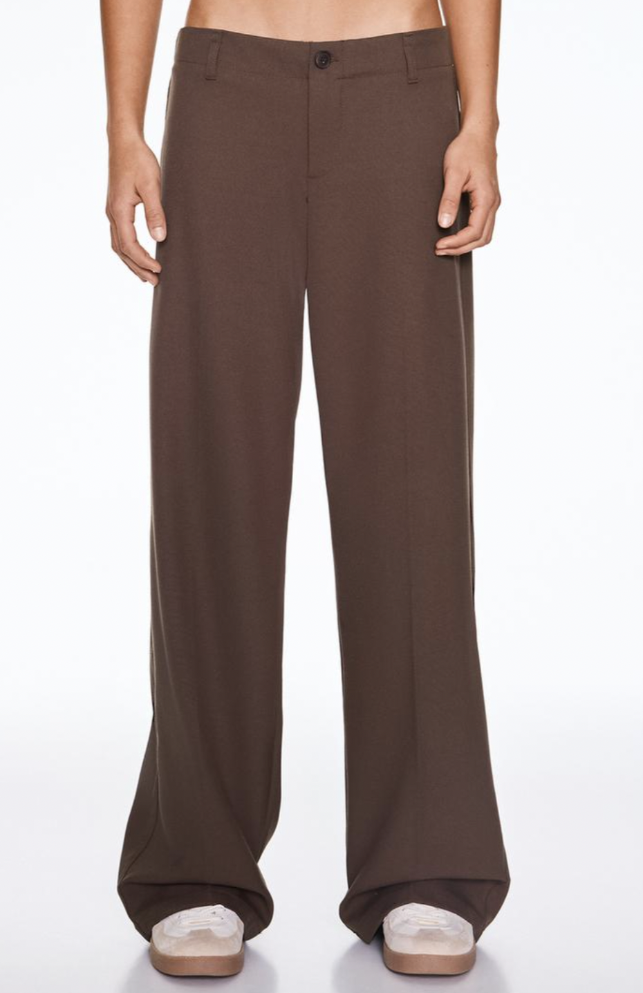 A person wearing brown wide-leg trousers stands against a plain white background. Their feet, in white shoes with thick soles, peek out at the bottom.