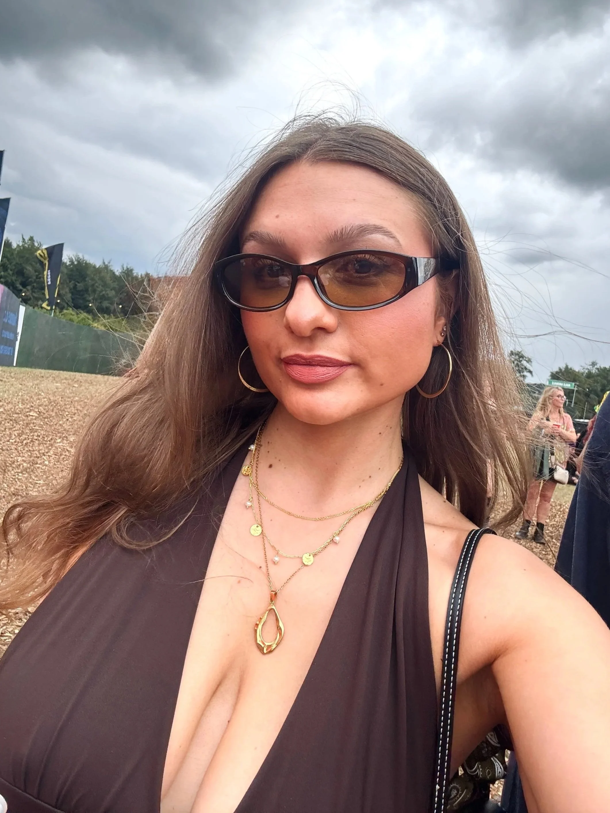 Selfie of a confident woman in sunglasses, a black halter top, and layered gold necklaces at an outdoor festival beneath stormy clouds.