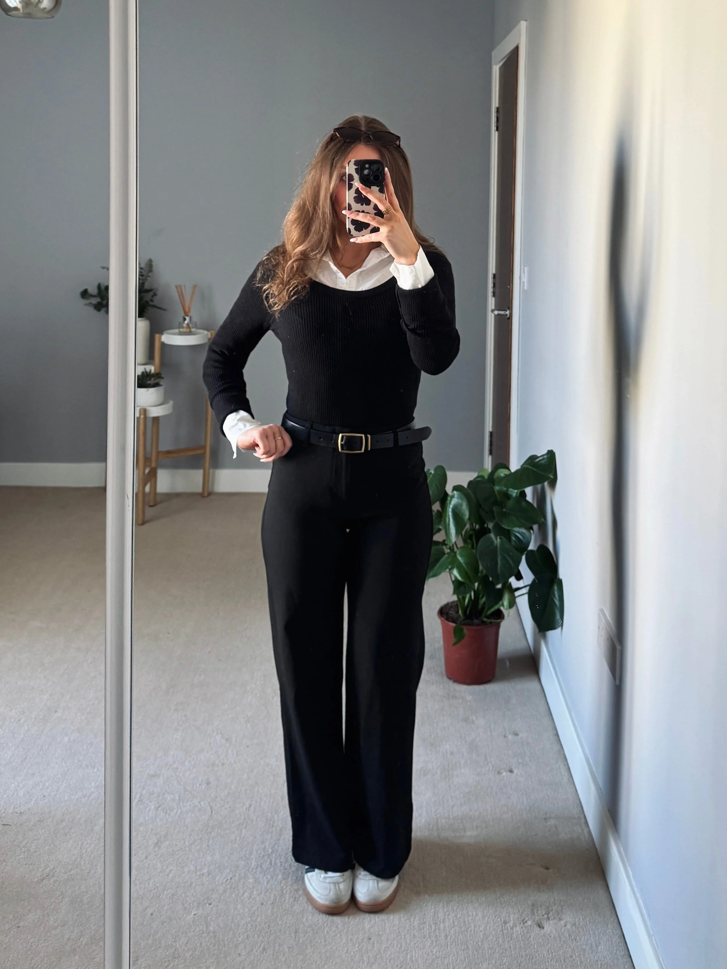 Woman taking a mirror selfie in a minimalist room. She wears a black sweater over a white shirt, black pants, and white sneakers. A potted plant is visible in the background.