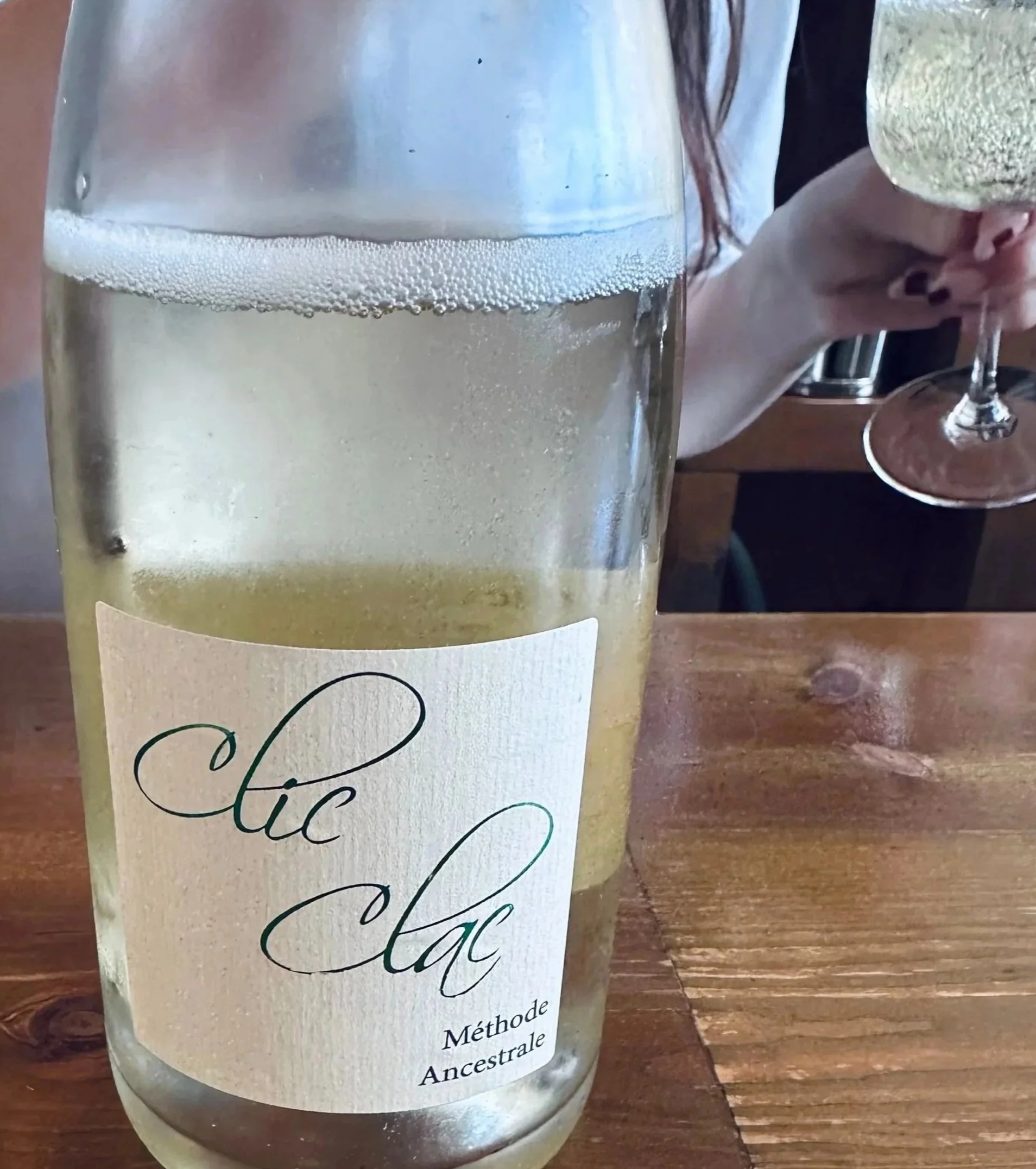 A bottle of "Clic Clac" Méthode Ancestrale sparkling wine sits on a wooden table. A person holds a glass filled with the bubbly drink, creating a casual, refined ambiance.