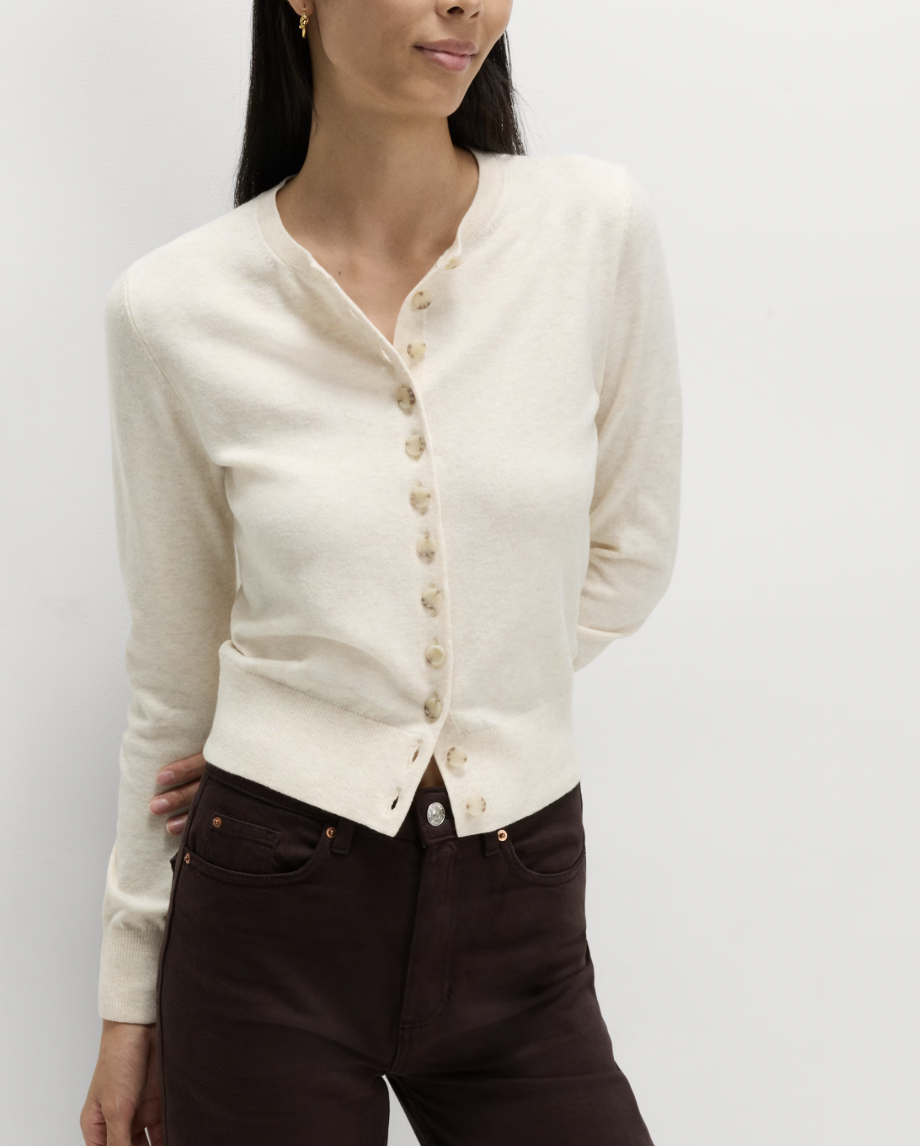 A woman in a cream cardigan with button details and dark pants stands against a white background, expressing a calm and relaxed demeanor.