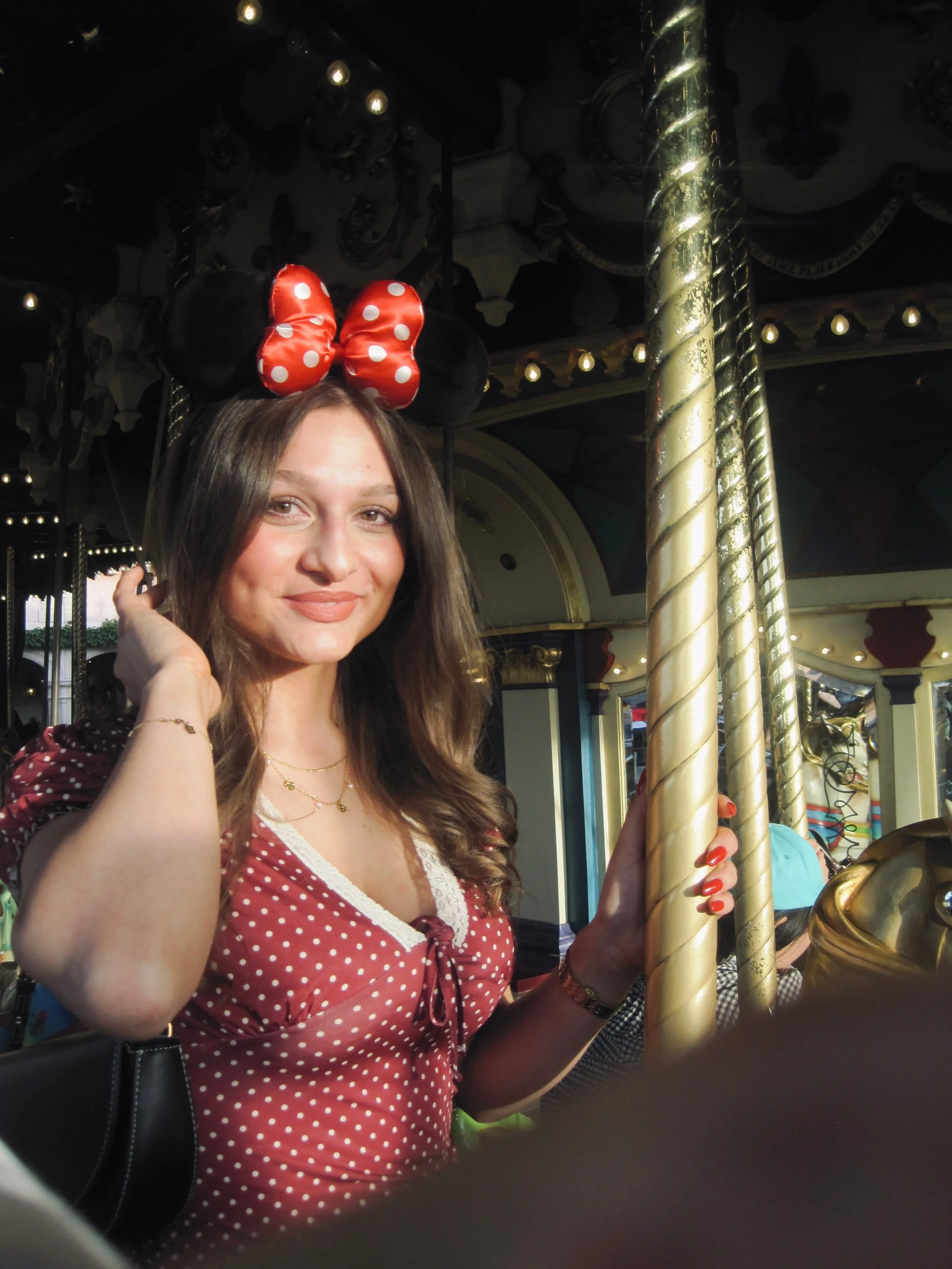 A smiling woman in a red polka-dot dress and Minnie Mouse ears poses beside a gold carousel pole, creating a cheerful theme-park mood.