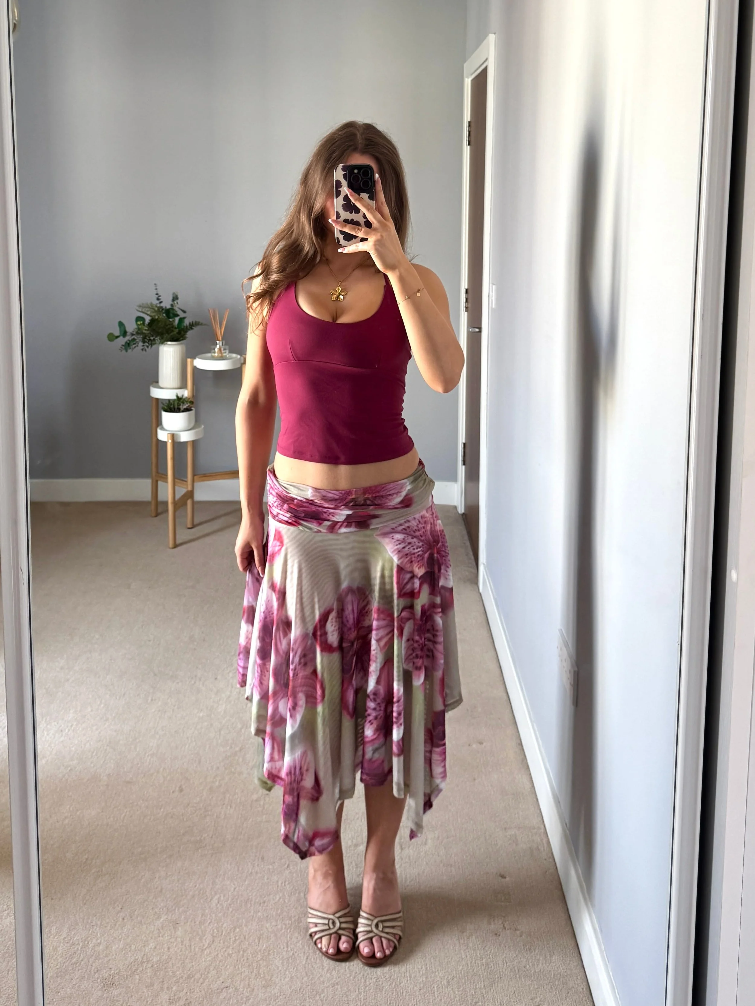 Woman taking a mirror selfie wearing a pink tank top, floral handkerchief skirt, gold necklace, and open-toe sandals in a minimalist room.