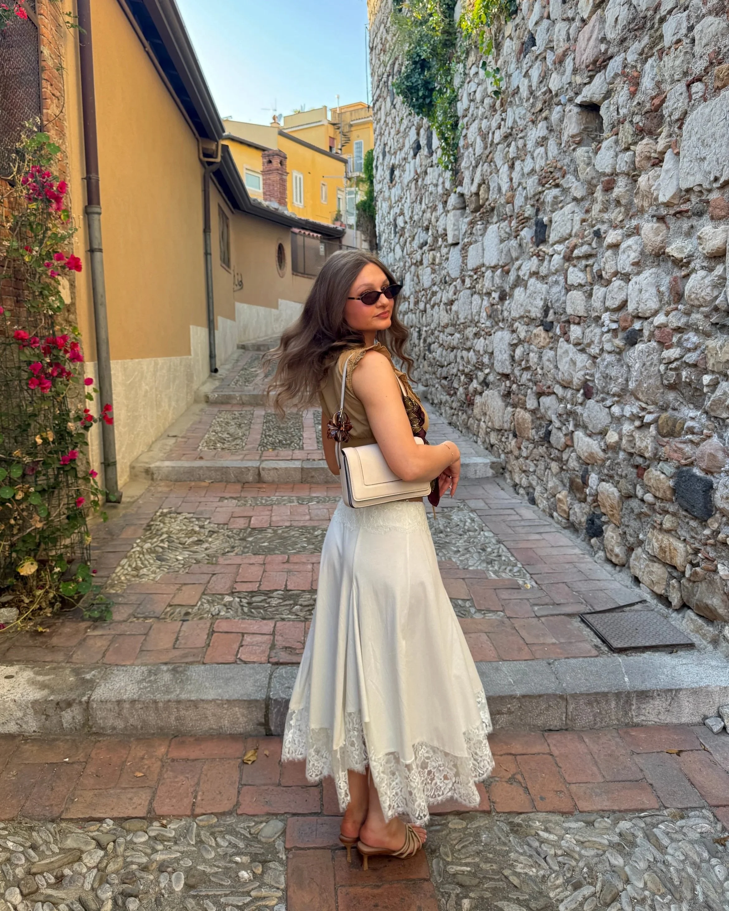 Young woman in sunglasses and white skirt posing on a narrow cobblestone street with stone and plaster walls and flowering plants.