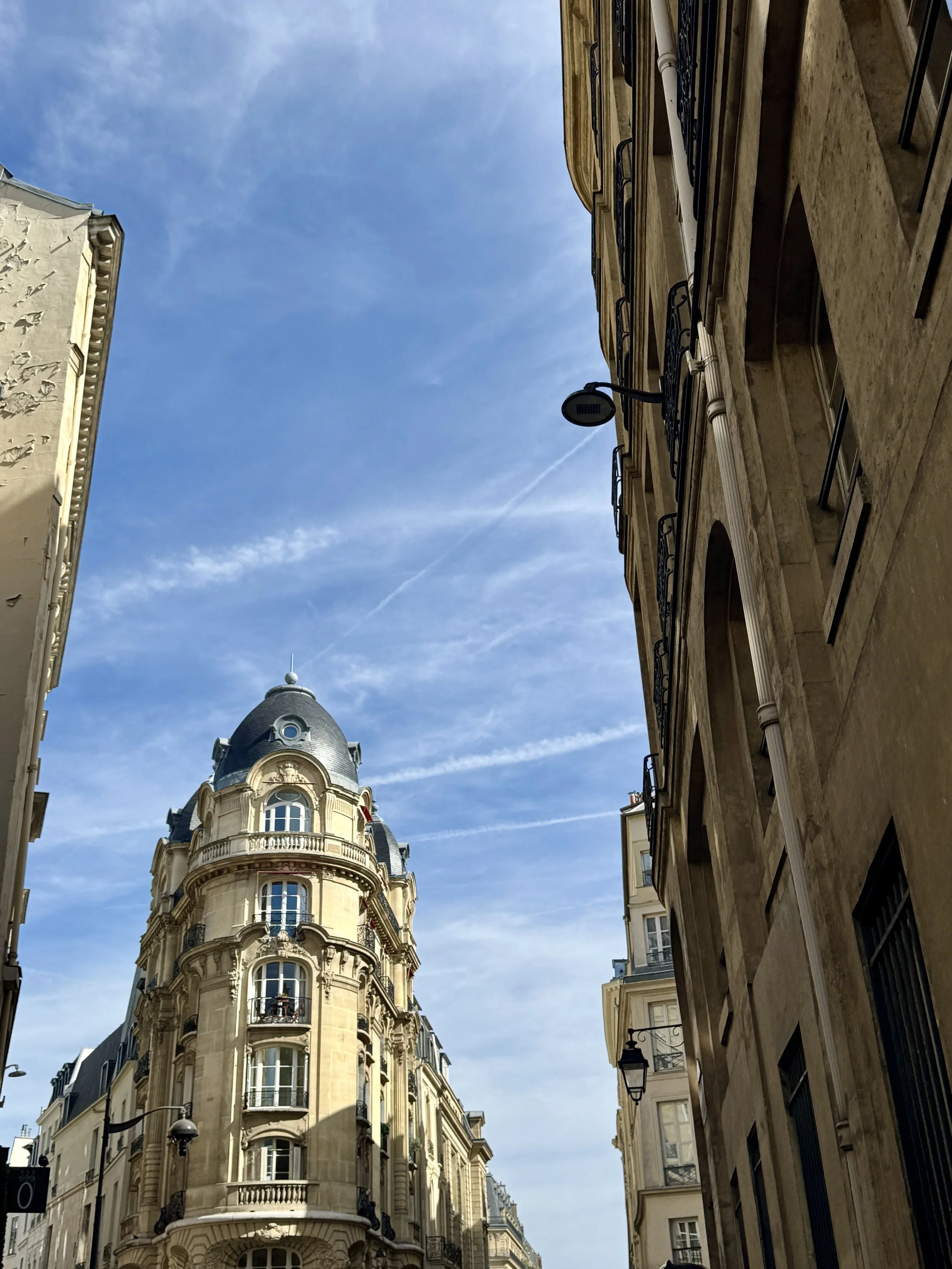 Looking up a narrow European street toward an ornate cream stone building with a domed roof, framed by tall façades beneath a bright blue sky.