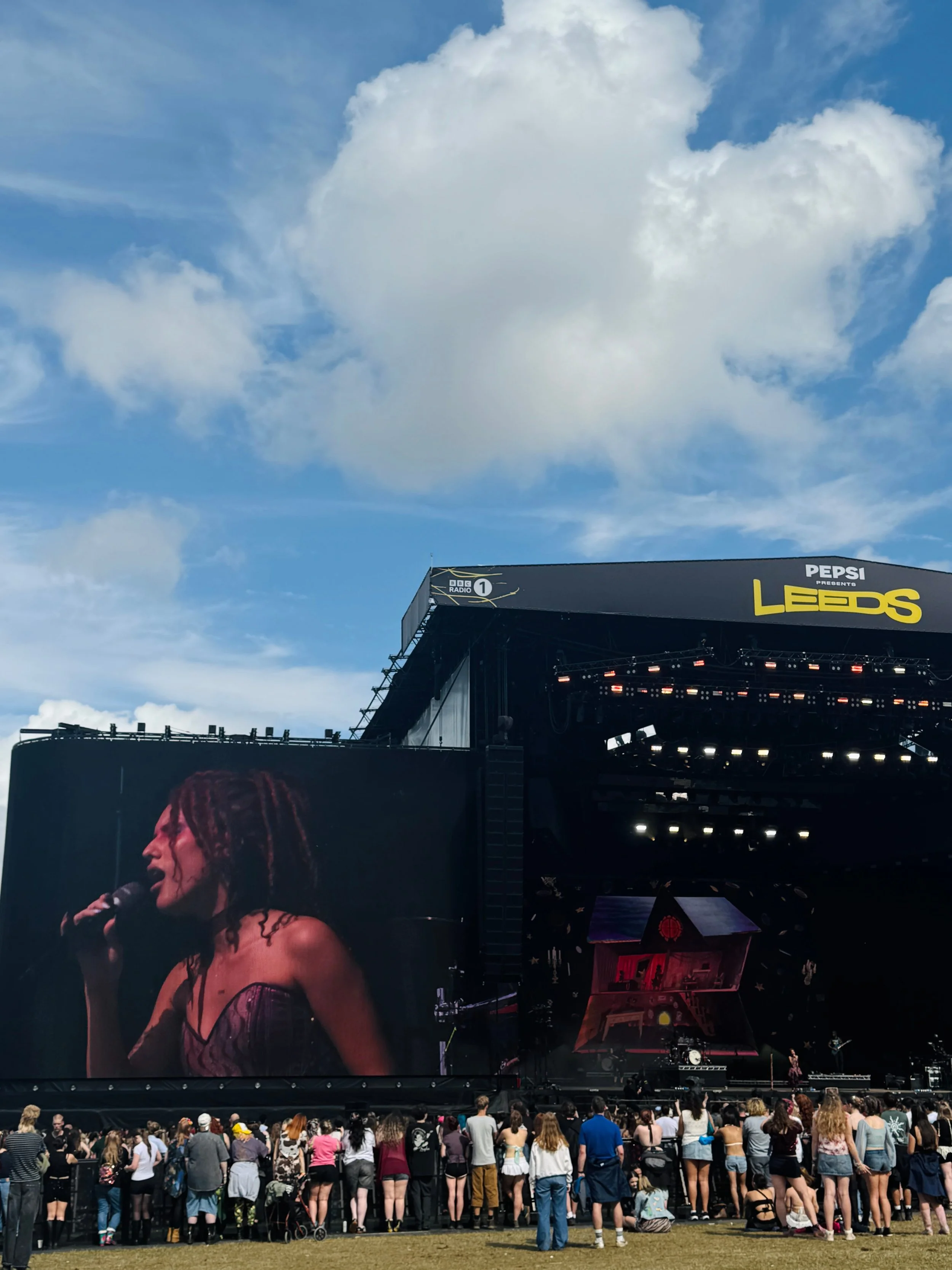 Outdoor Leeds music festival stage with Absolutely performing on a giant screen, cheering crowd below, and dramatic white clouds in a blue sky.