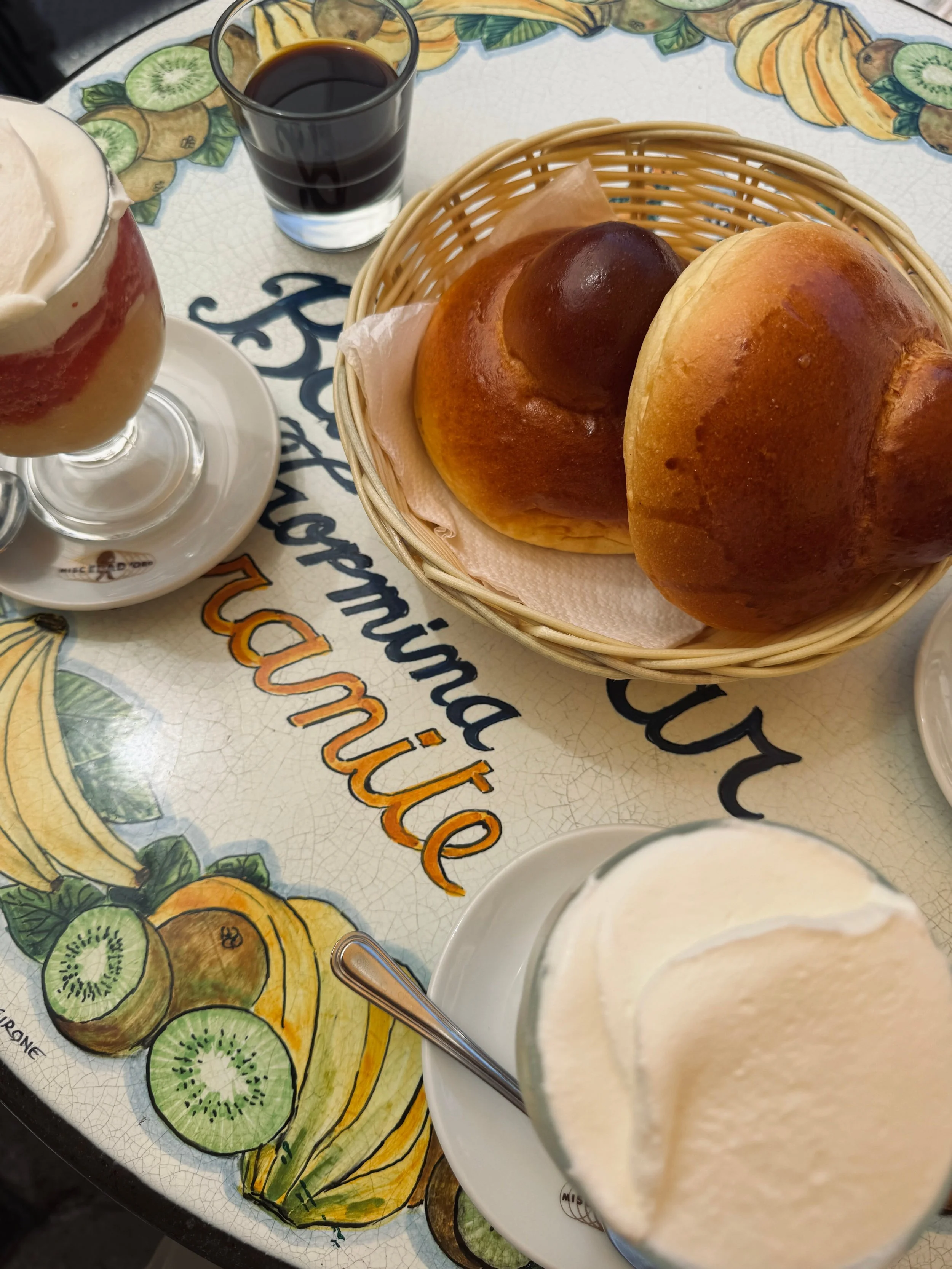 Table with two brioche buns in a basket, two glasses of gelato with spoons, and a small glass of dark espresso.