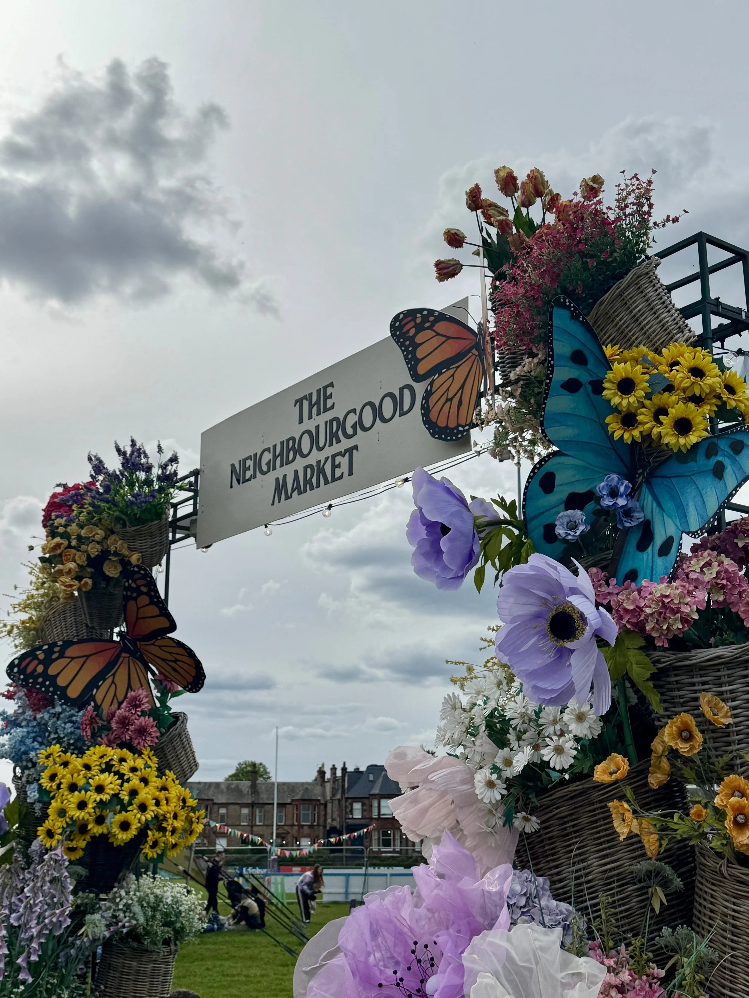 Floral arch with butterflies and "The Neighbourgood Market" sign under a cloudy sky. Vibrant flowers create a lively and inviting atmosphere.