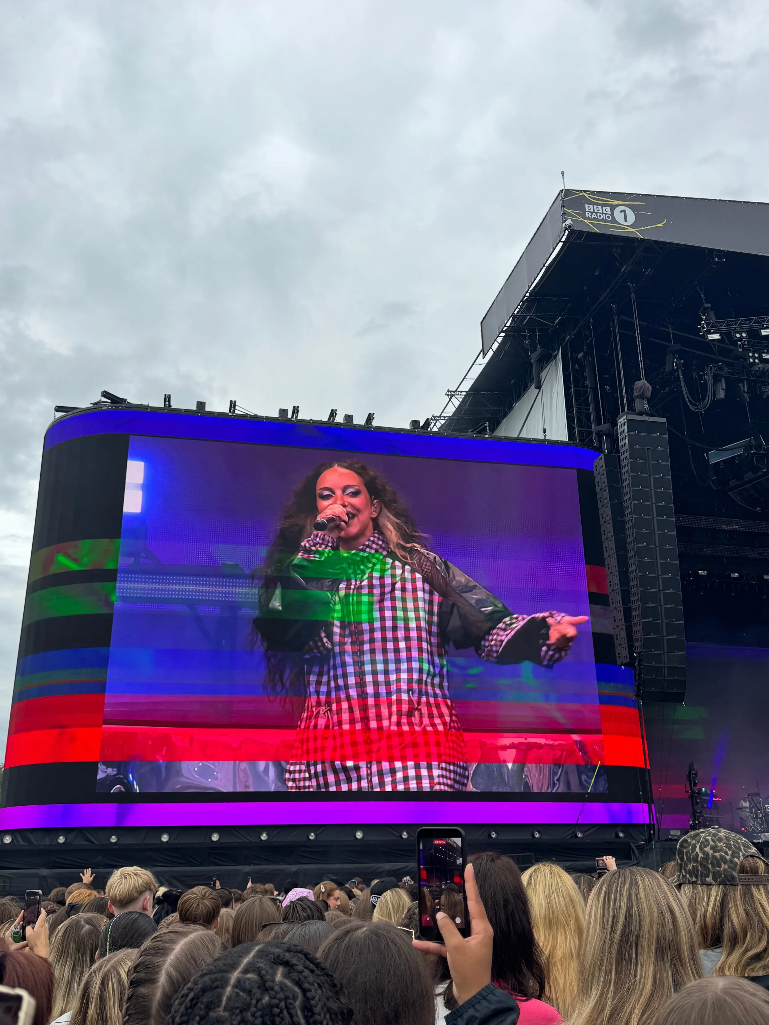 JADE performs energetically on a giant stage screen with colorful glitch effects as a dense crowd records the concert under cloudy skies.