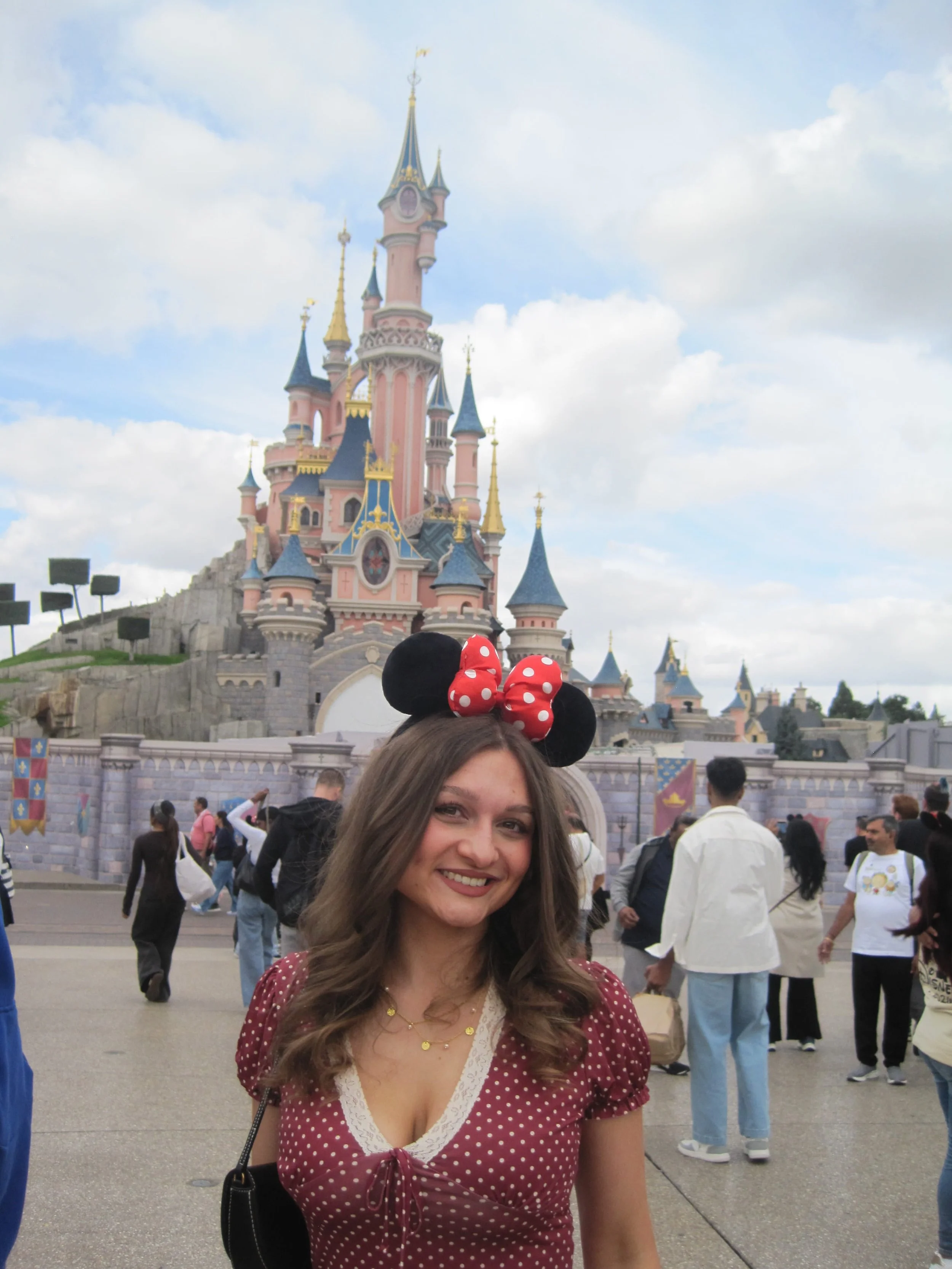 A smiling woman in Minnie Mouse ears poses before a fairytale castle at a busy theme park, creating a cheerful, magical vacation scene.