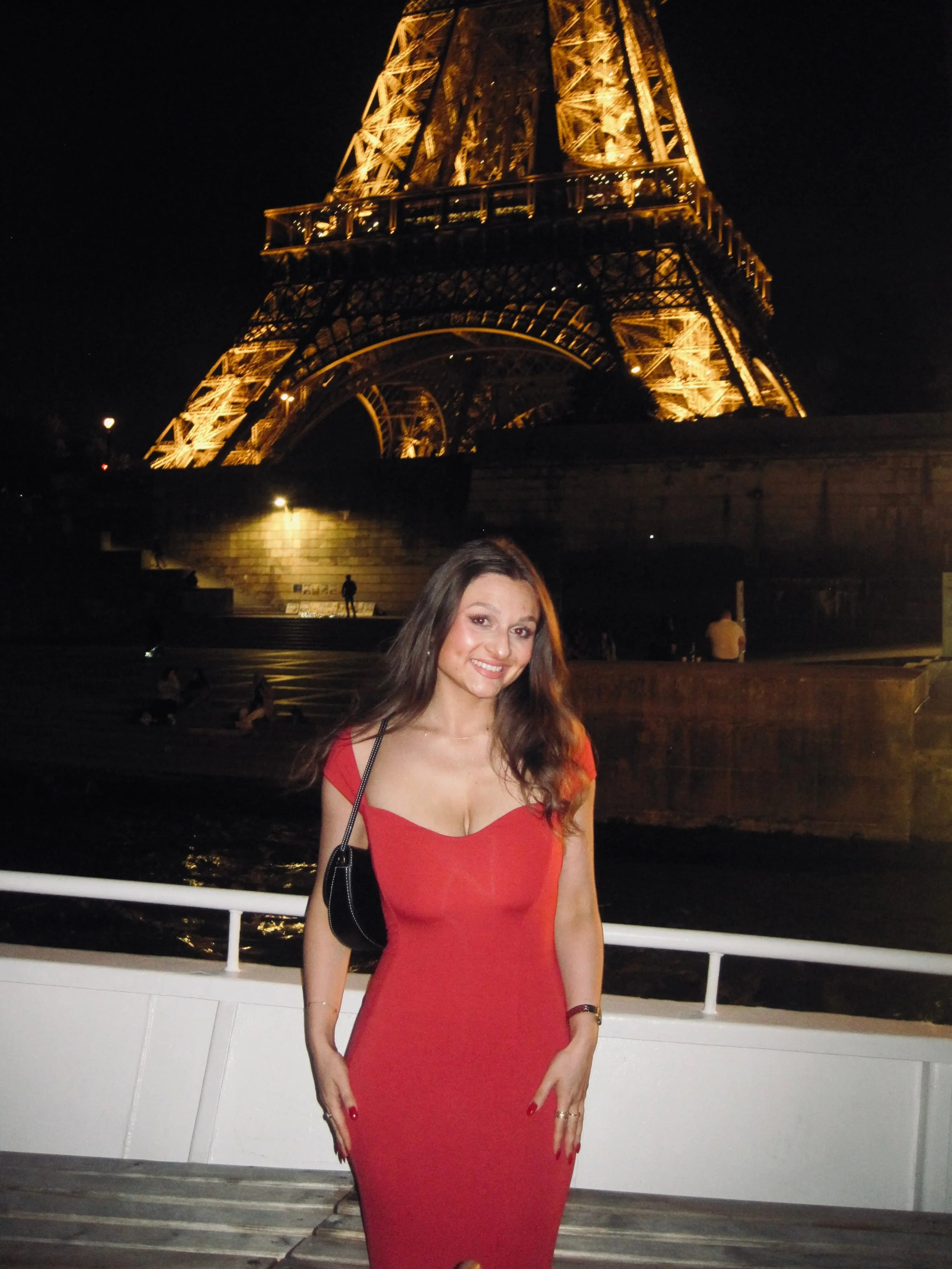Smiling woman in a red dress poses on a boat at night, with the warmly illuminated Eiffel Tower rising behind her against a dark sky.