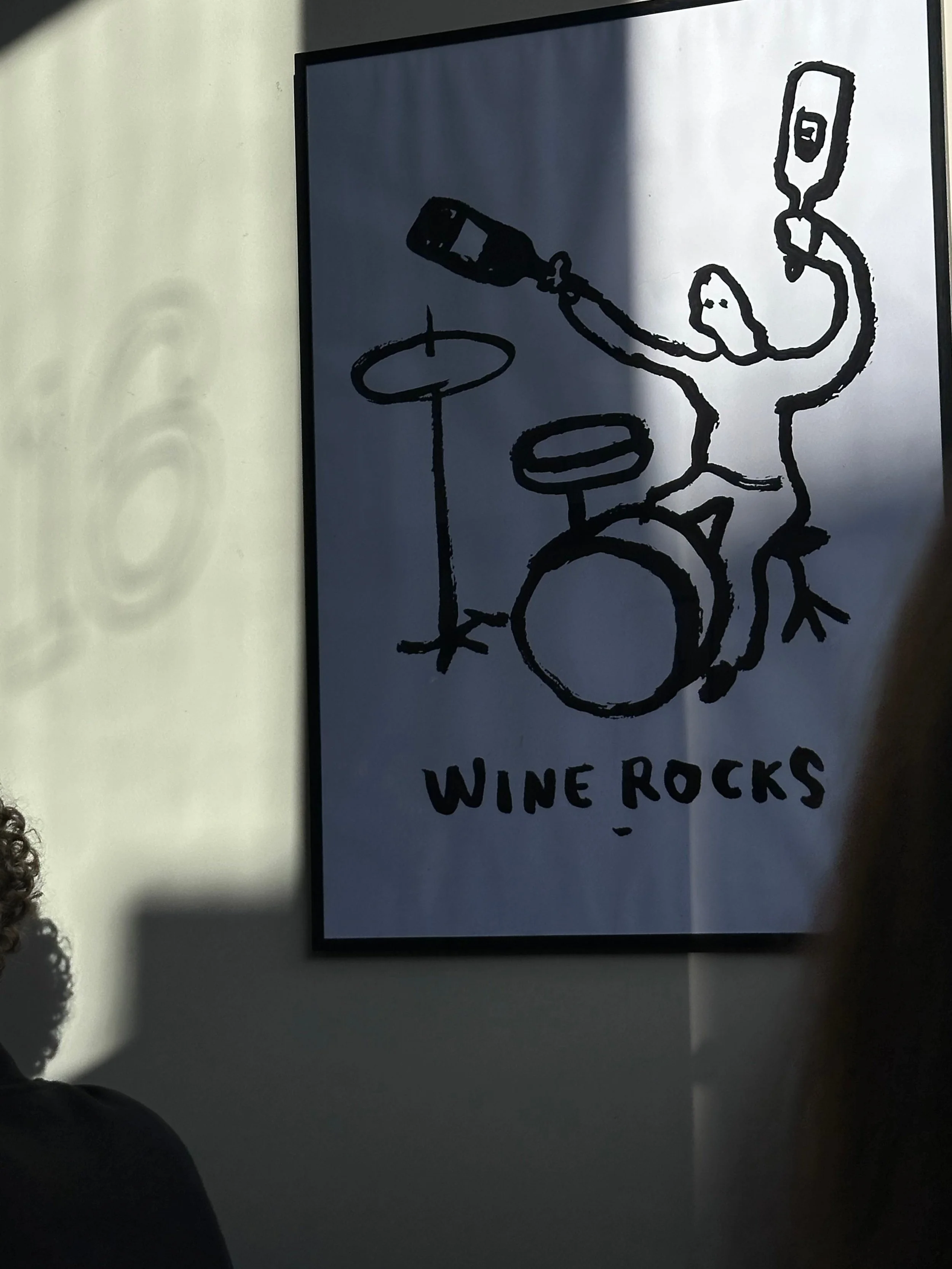 A framed poster features a stick figure playing drums with wine bottles, accompanied by the text "Wine Rocks." The scene is light-hearted and playful. Shadows fall across the wall.
