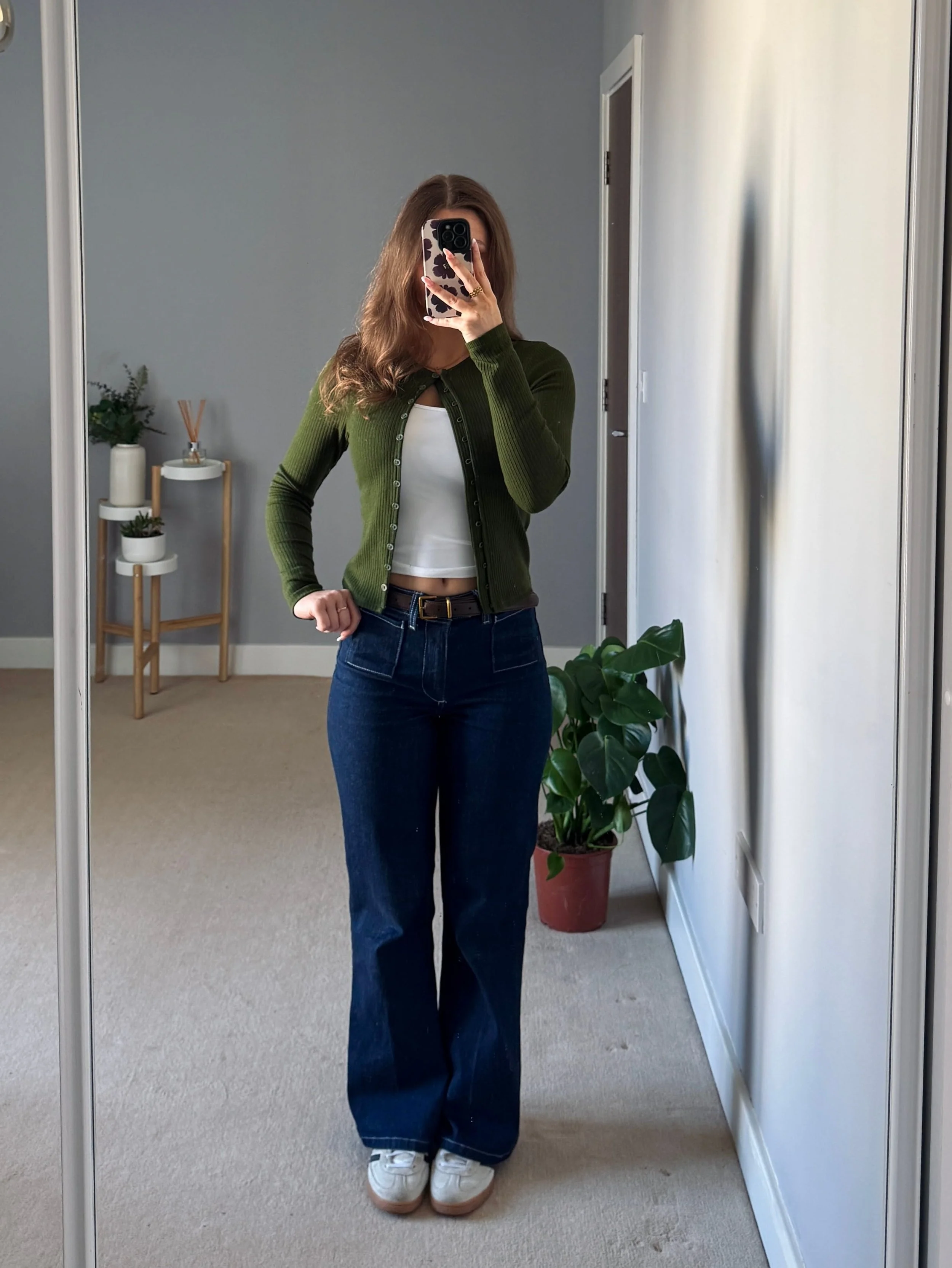 A woman takes a mirror selfie in a cozy room, wearing a green cardigan, white top, and blue jeans. A potted plant and decor are in the background.