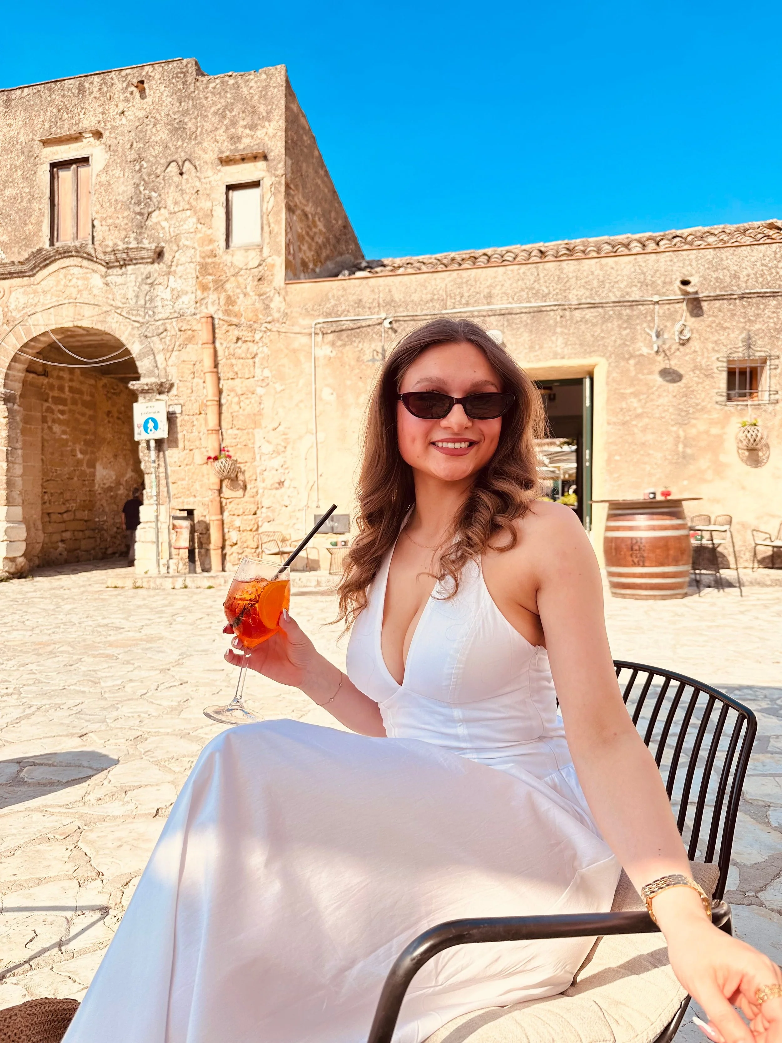 A woman in a flowing white dress sits elegantly on a chair, holding a refreshing drink in her hand.
