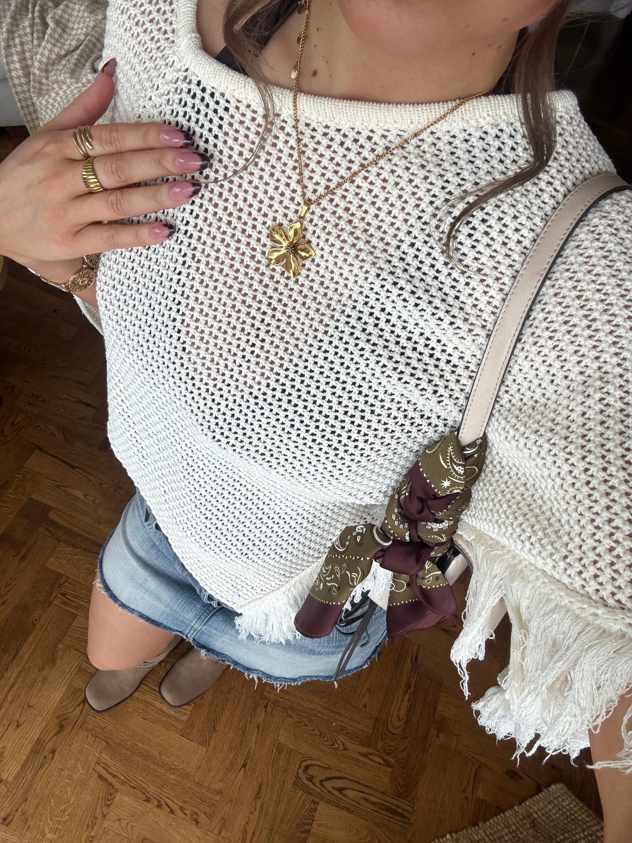 Woman wearing a white knit fringe sweater, denim shorts, brown boots, gold flower necklace, rings, pink manicure, and a beige handbag with a scarf tied to it.