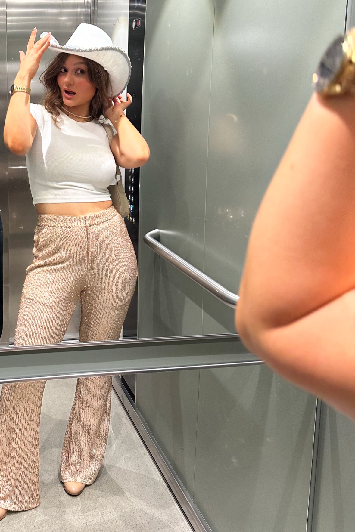A woman looking in a mirror wearing gold sequin trousers and a white cowboy hat