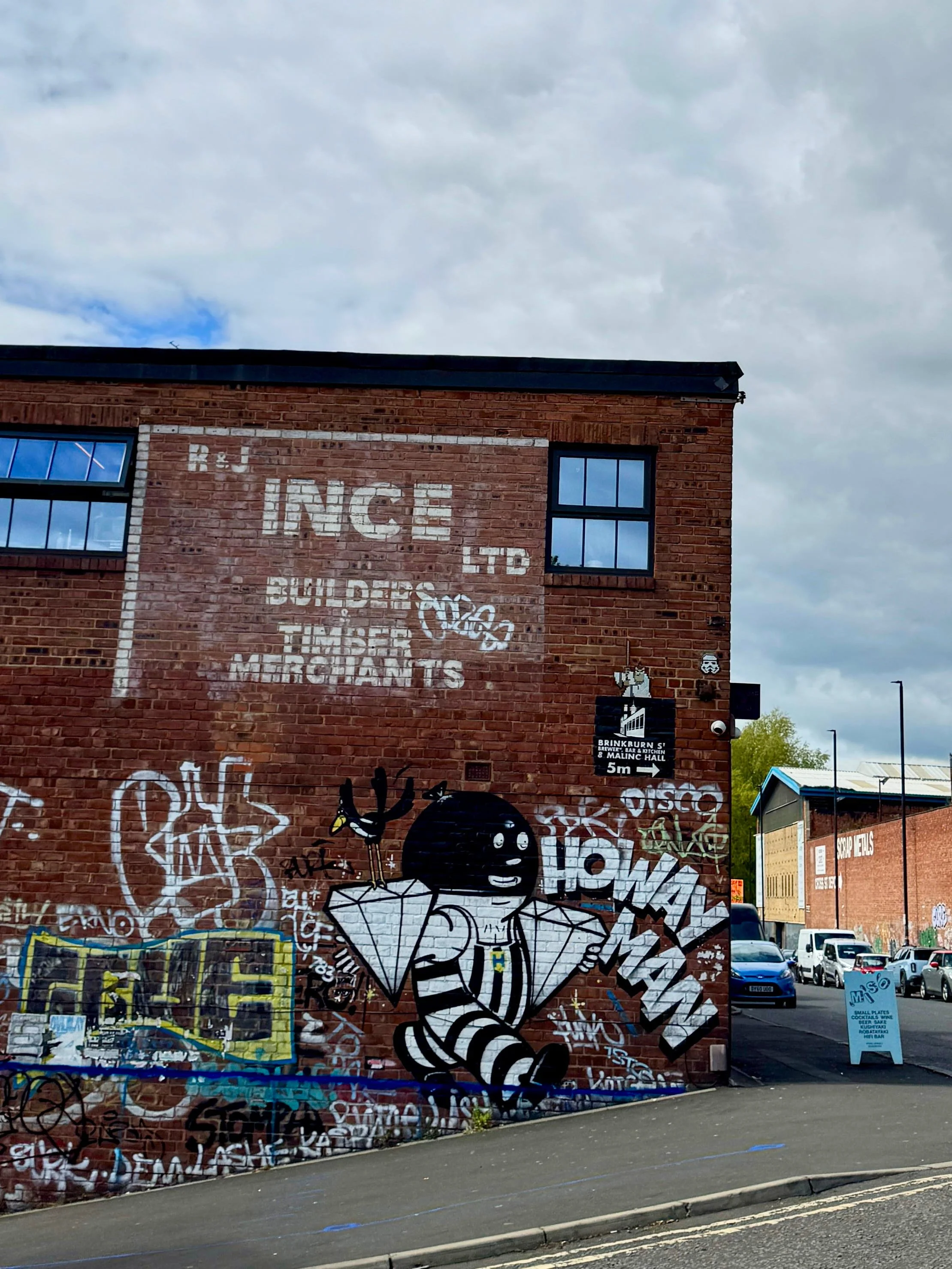 A brick building with faded white signage. Vibrant graffiti, including a cartoon bee, adorns the lower wall. Cloudy sky above conveys a moody urban vibe.