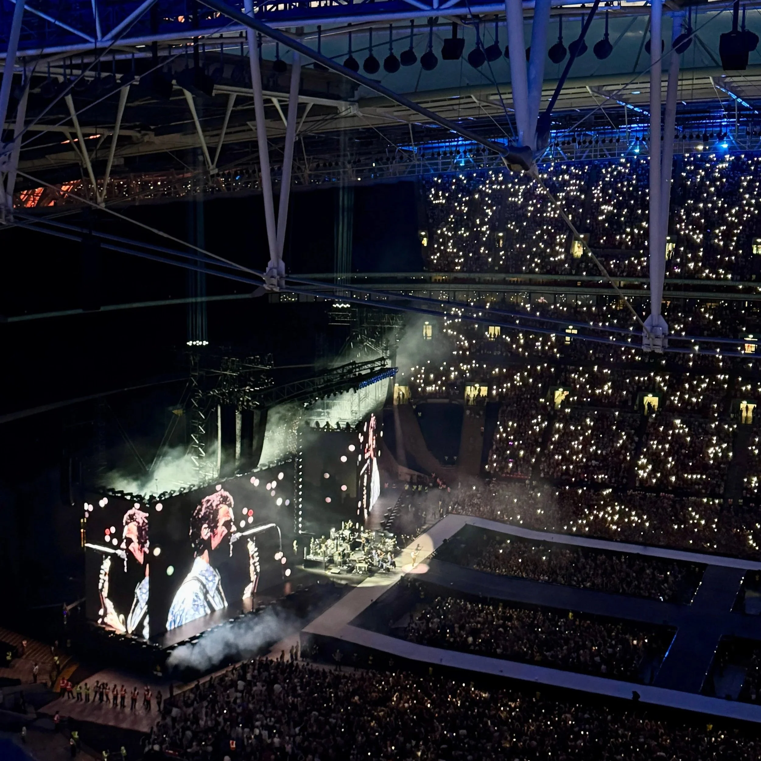 A bustling Wembley Stadium during a Harry Styles concert, with a sea of fans gathered around the illuminated stage.