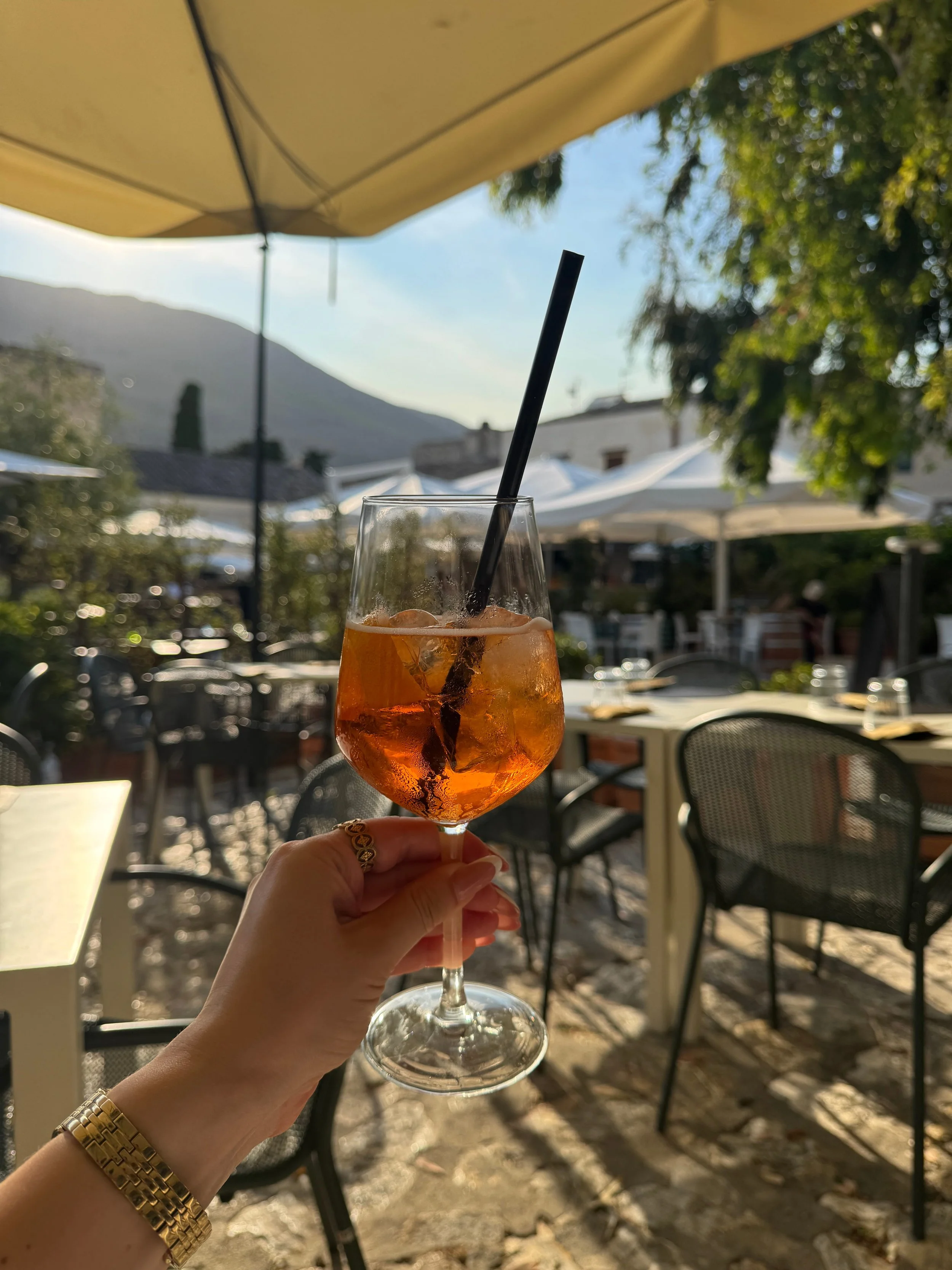 A person sits at an outdoor table, sipping a vibrant Aperol Spritz, with a lively atmosphere around them