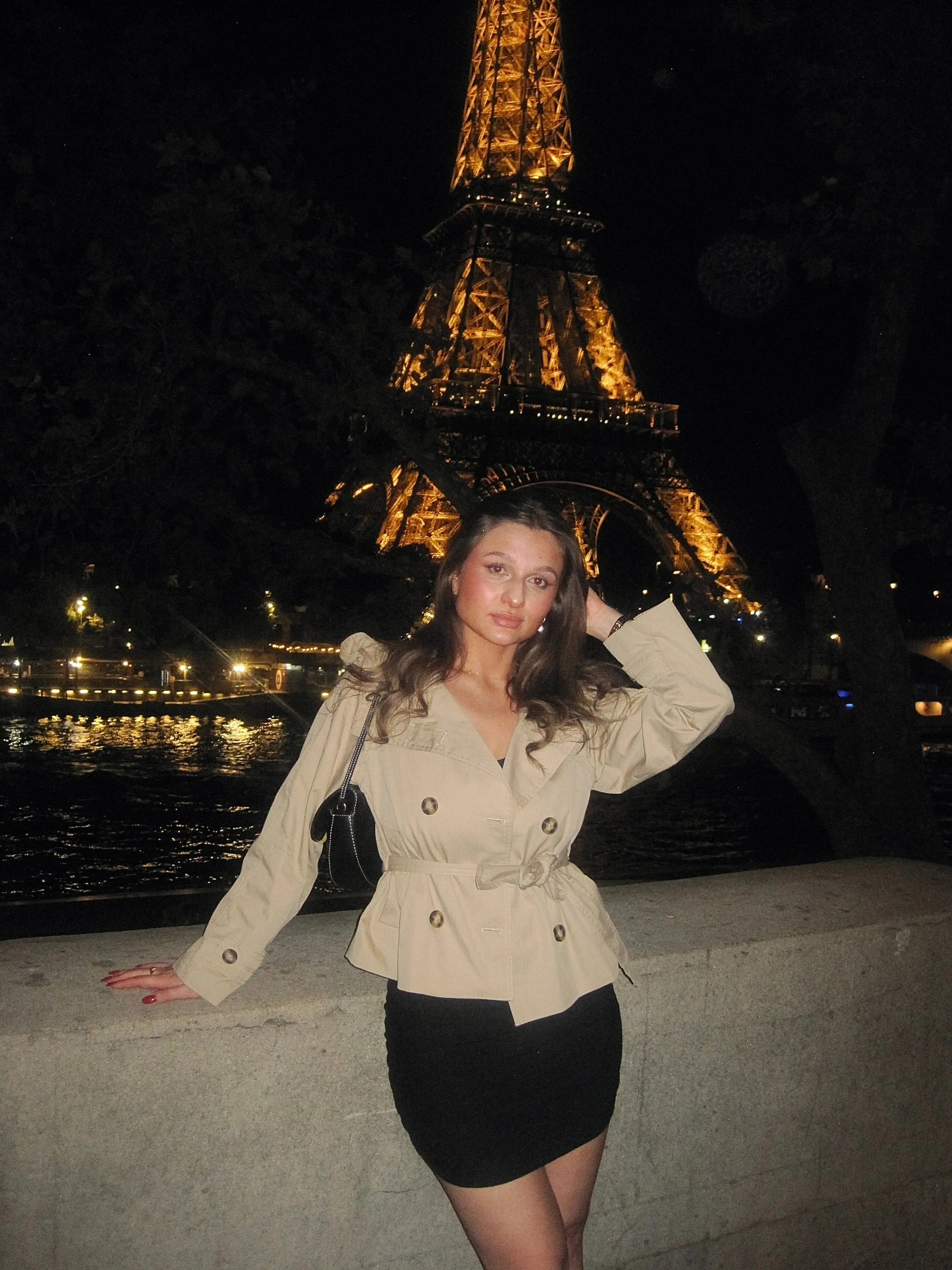 Woman in a beige coat poses by the Seine at night, with the warmly illuminated Eiffel Tower behind her, creating a romantic Parisian mood.