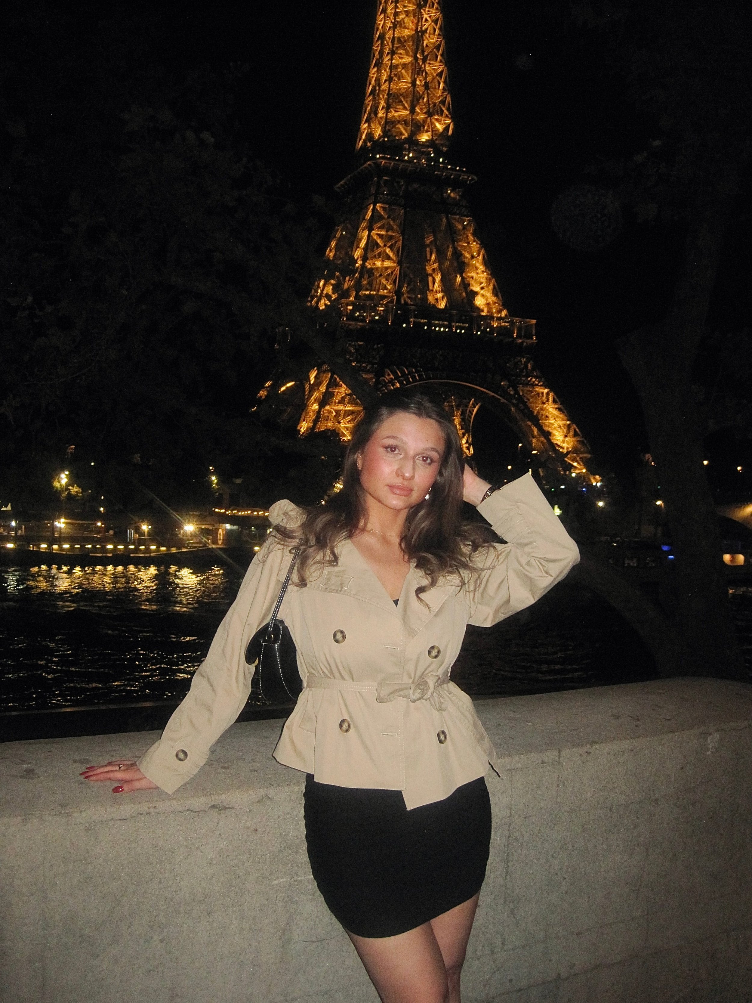 A woman in a beige coat and black skirt poses by the Seine at night, with the golden-lit Eiffel Tower behind her, creating a romantic Paris scene.