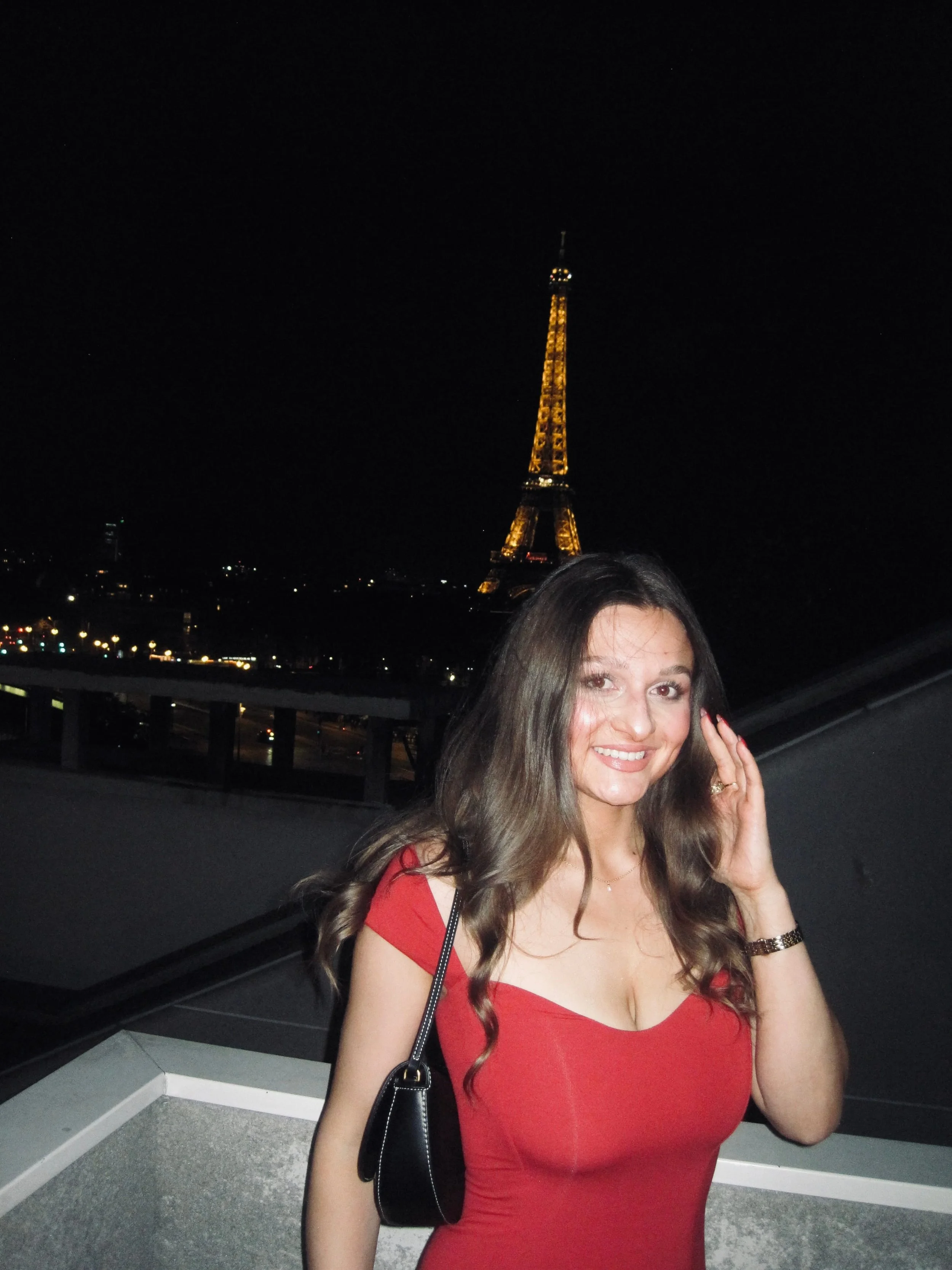 Smiling woman in a red dress poses on a rooftop at night, with the illuminated Eiffel Tower and twinkling Paris city lights behind her.