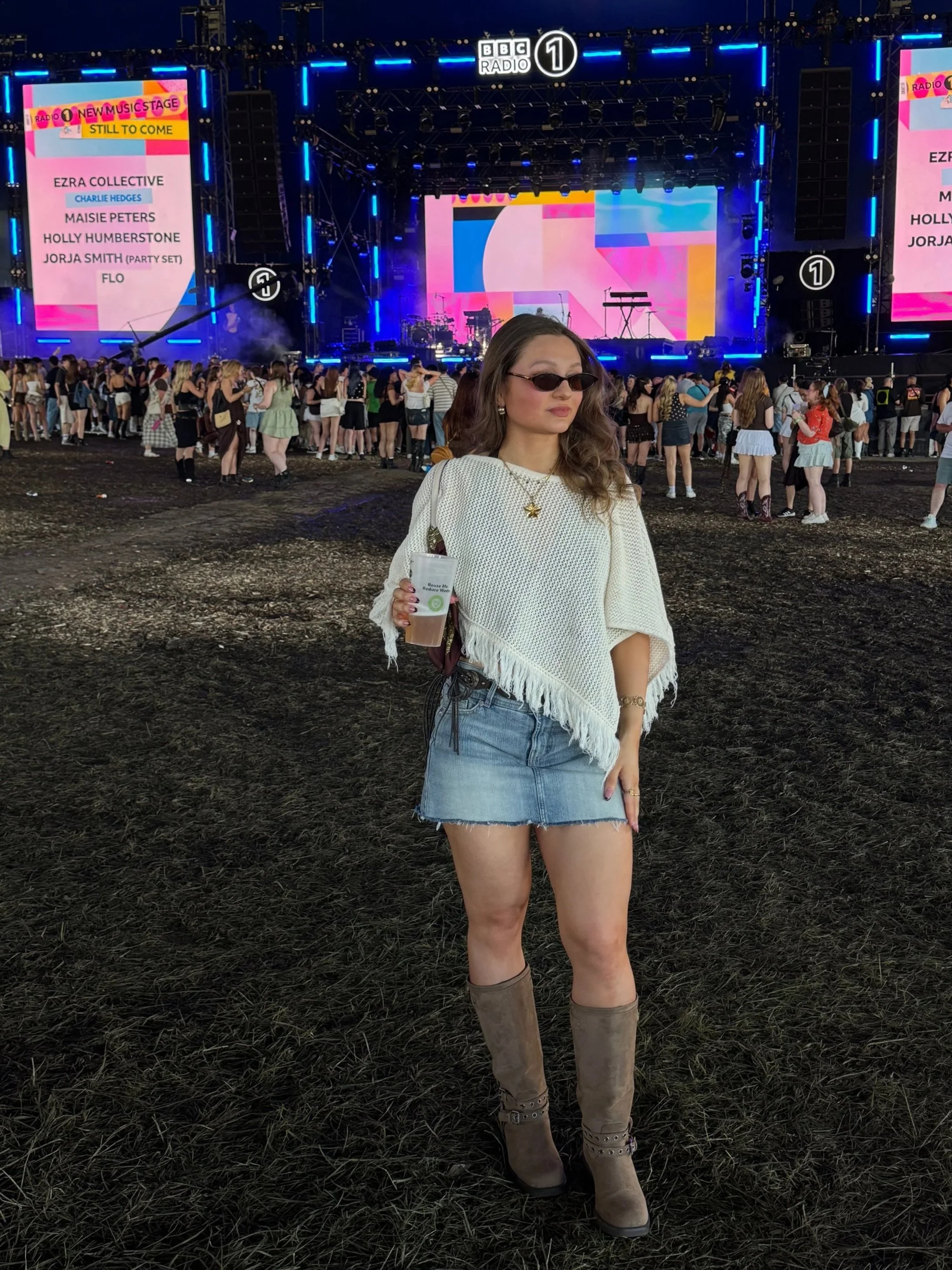 A woman in sunglasses, a white poncho, denim skirt, and boots stands holding a drink at an outdoor music festival with a large stage and crowd in the background.