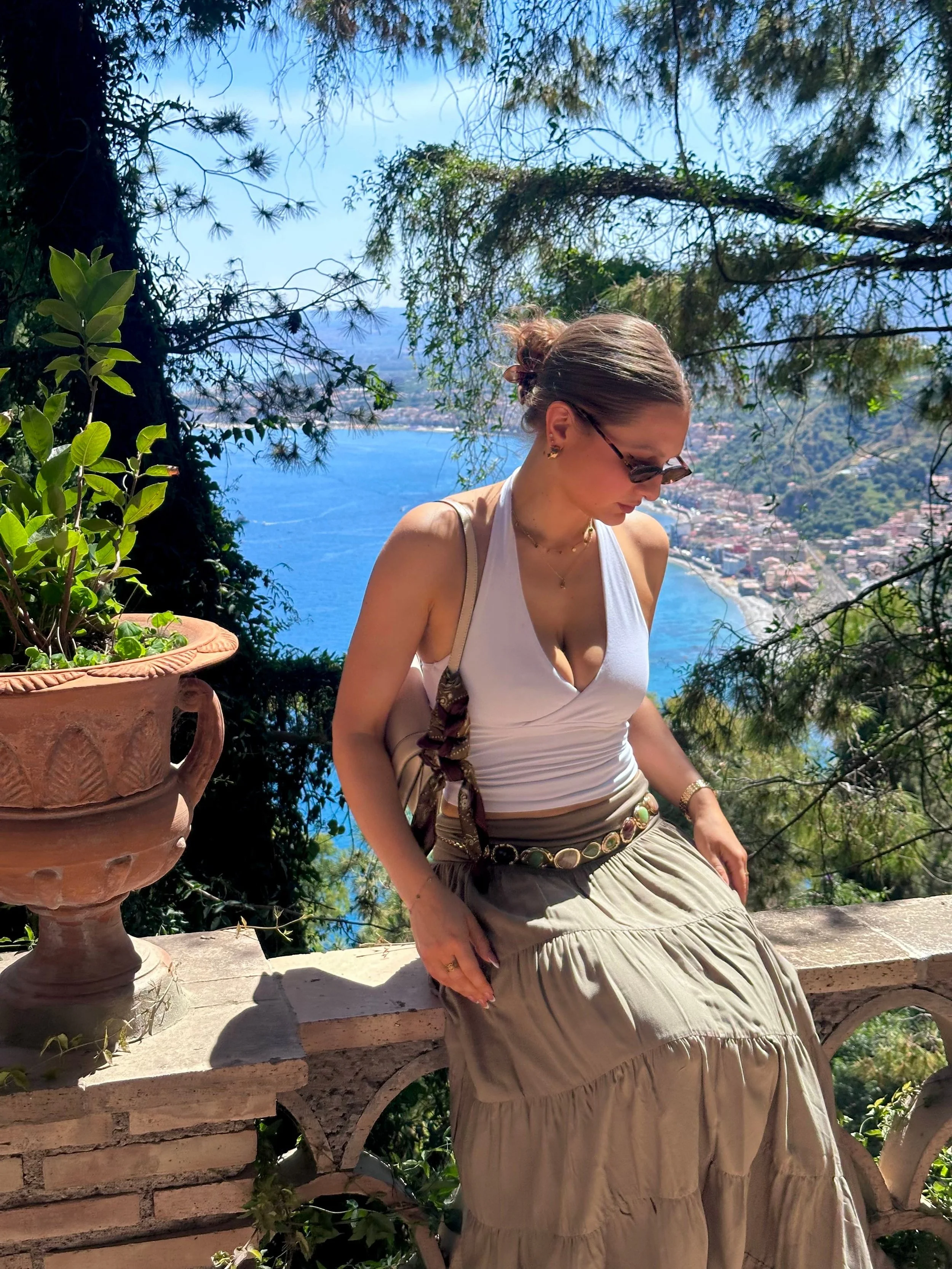 Woman in a white top and beige skirt sitting on a stone railing with a scenic coastal view and greenery in the background.